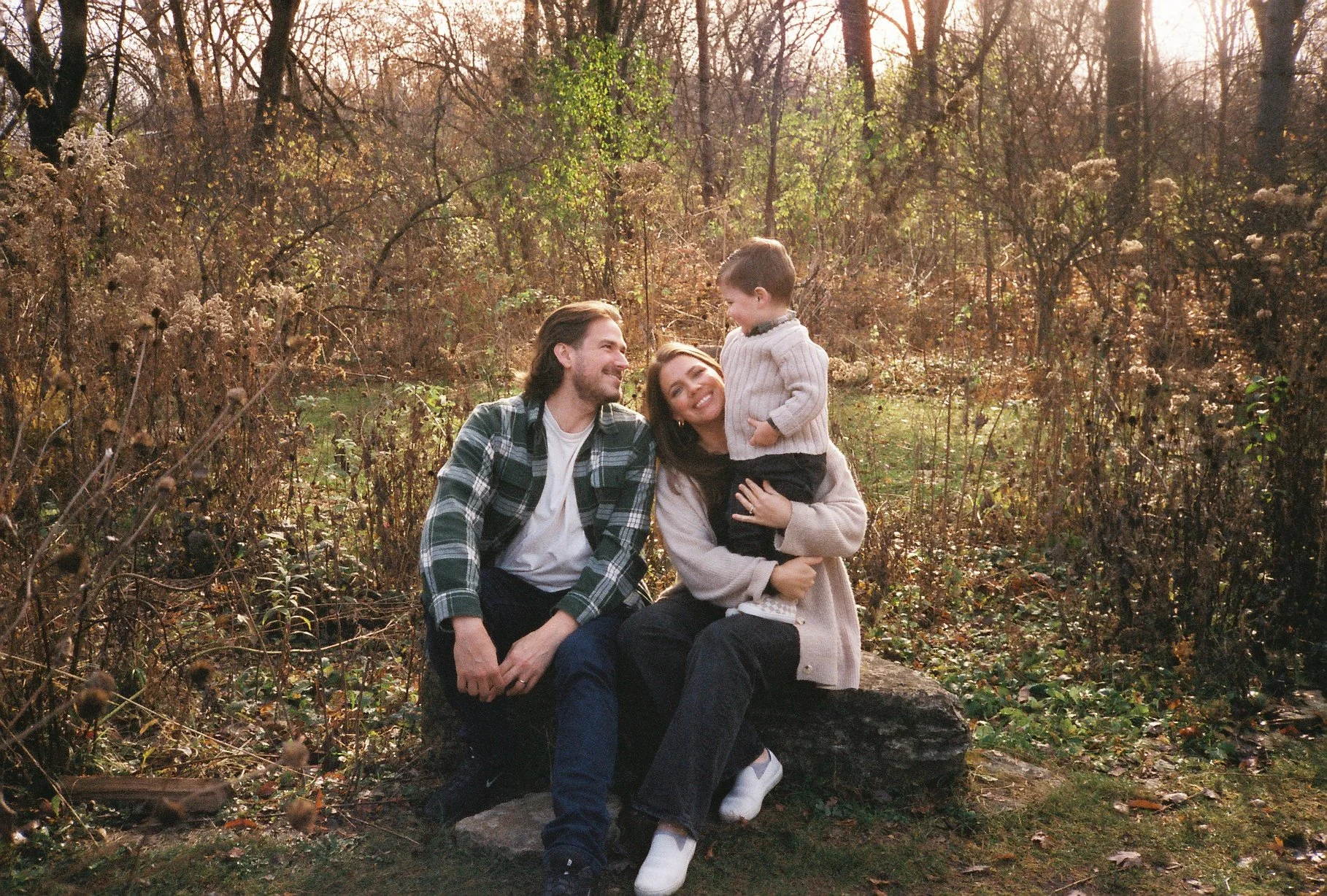 A family of three sitting on a log in a wooded area during sunset, smiling and enjoying each other's company.