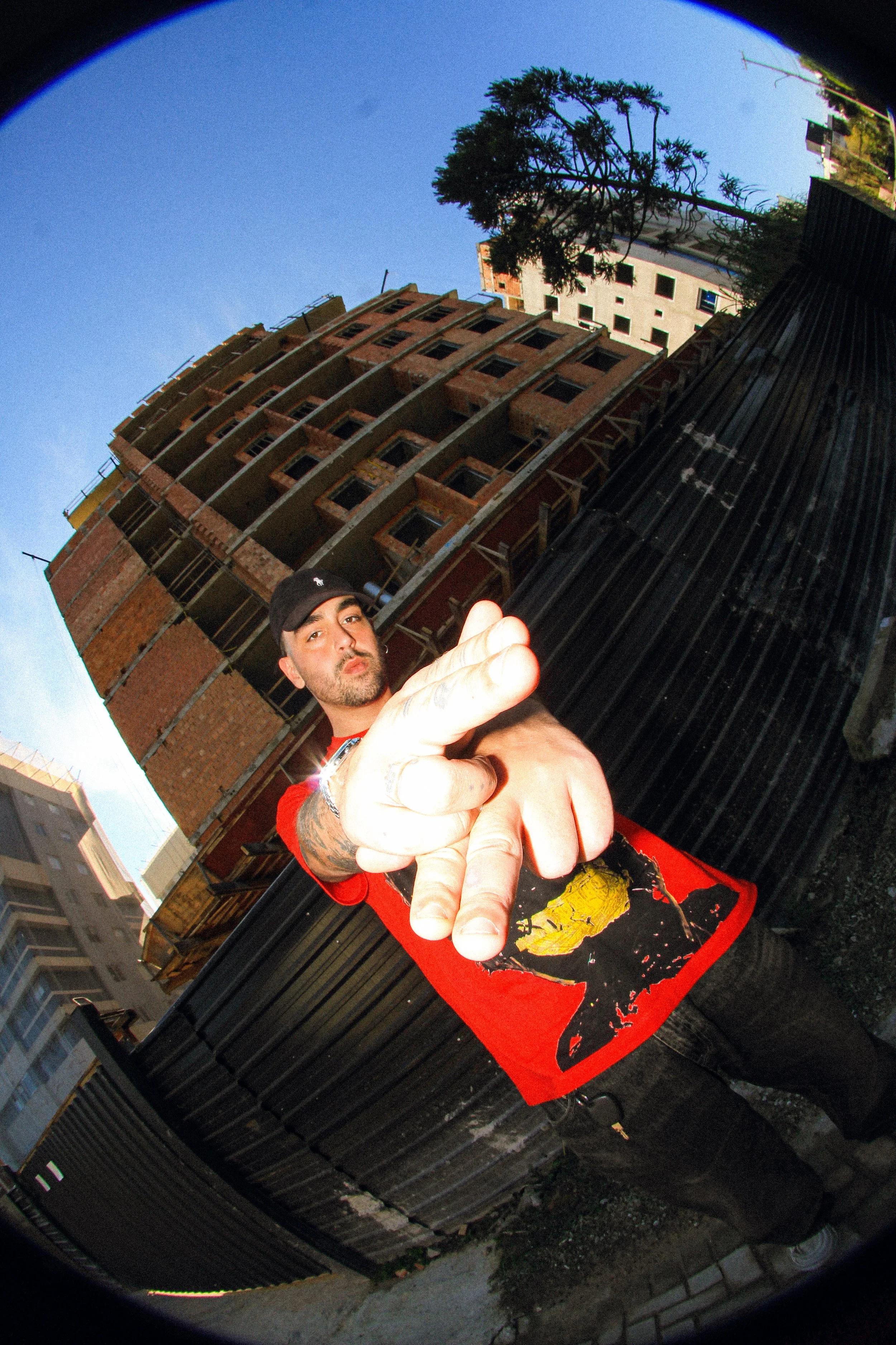 A man in a red shirt with a graphic print, black pants, and a black cap stands in front of a black fence and a multi-story building under construction, making a hand gesture towards the camera.