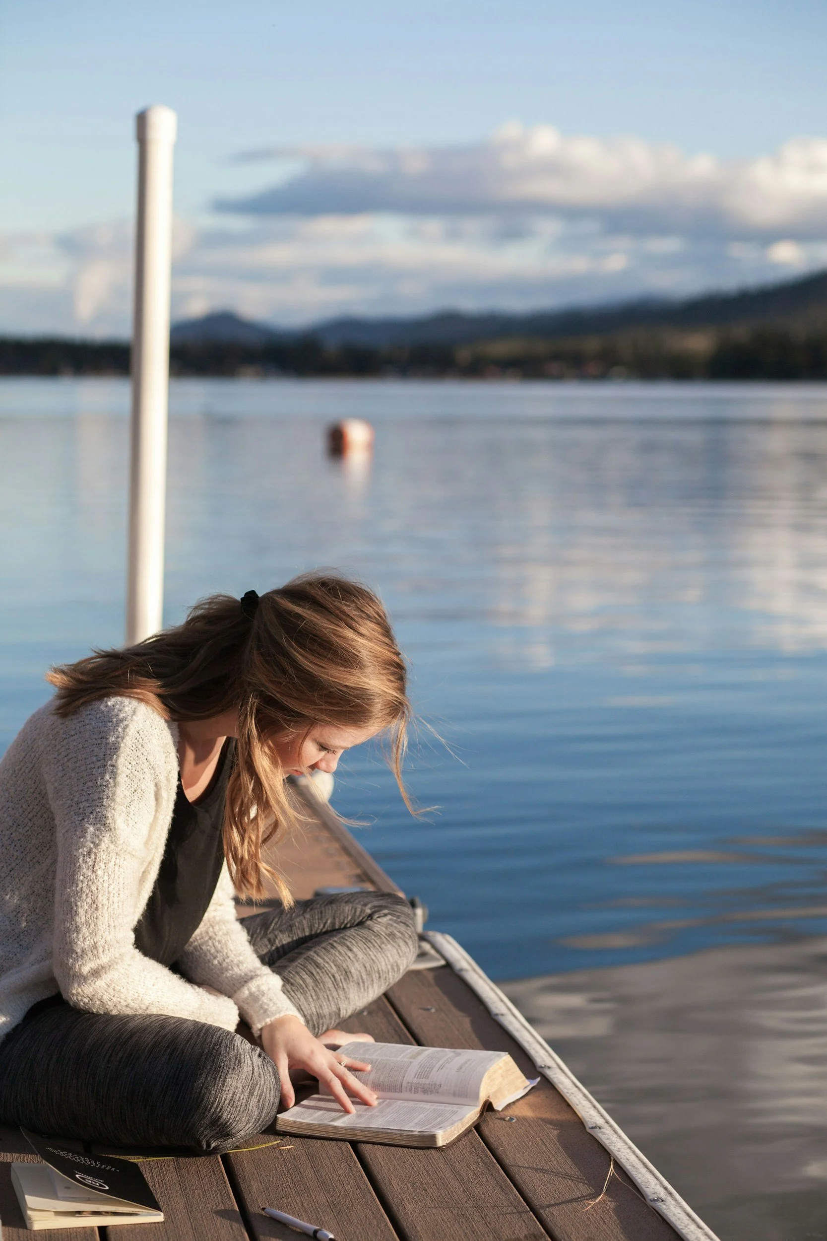 A teenage girl sitting on a waterfront dock reading a book