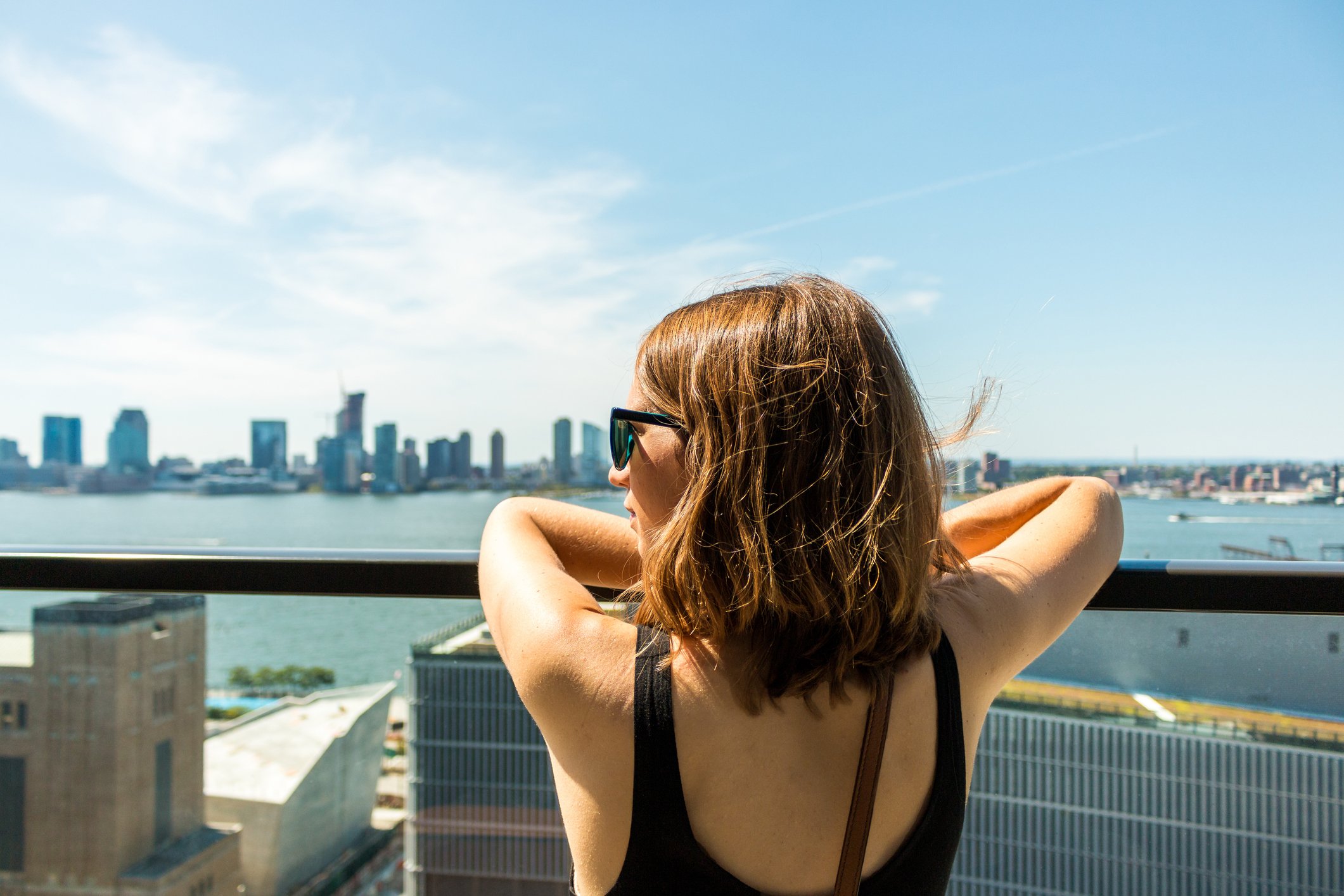 A woman with brown hair and sunglasses leaning on a balcony railing, looking at the NYC skyline view in the distance