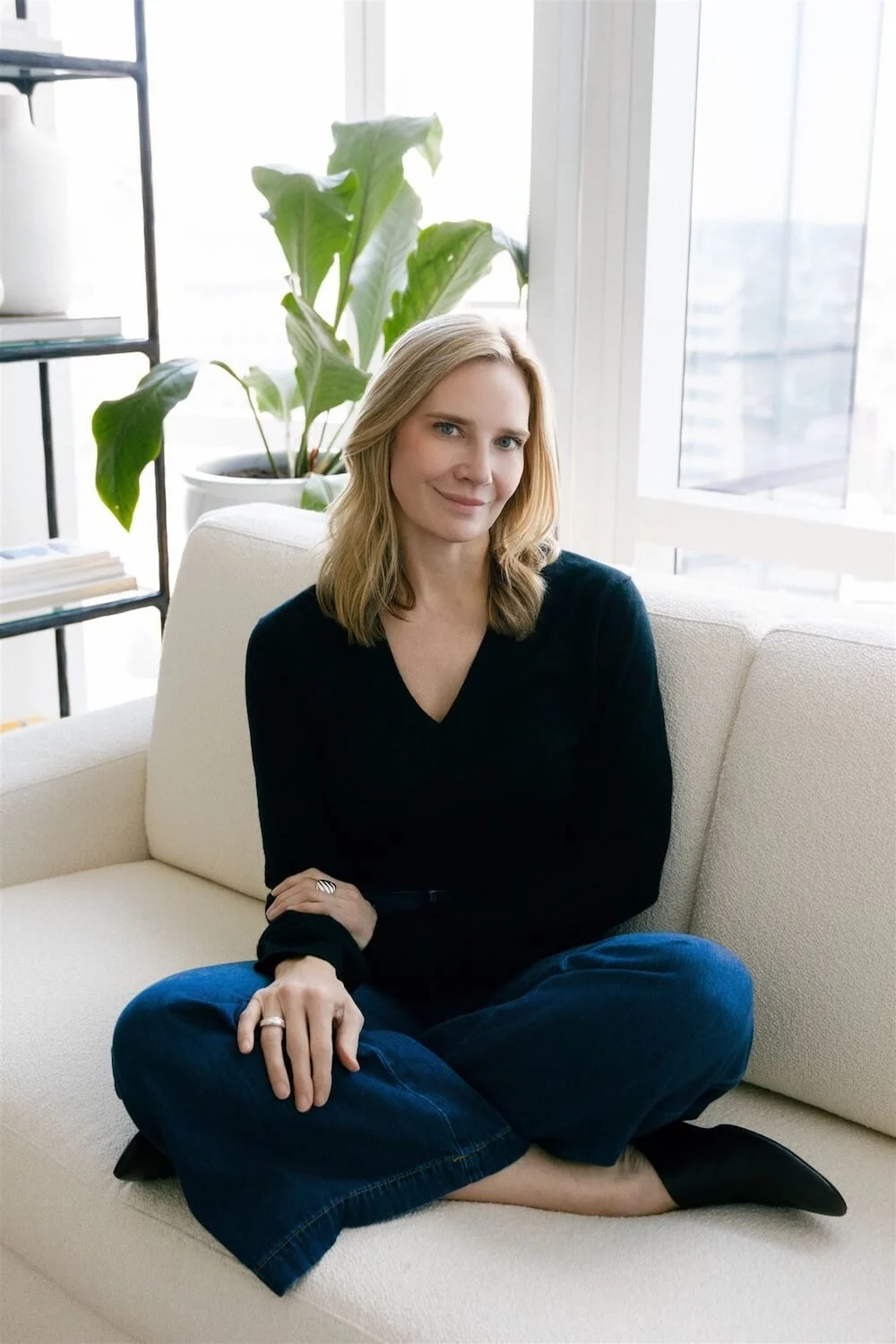 Therapist Jennifer Warpool sits cross-legged on a cream couch, wearing dark jeans and a navy sweater, smiling empathetically
