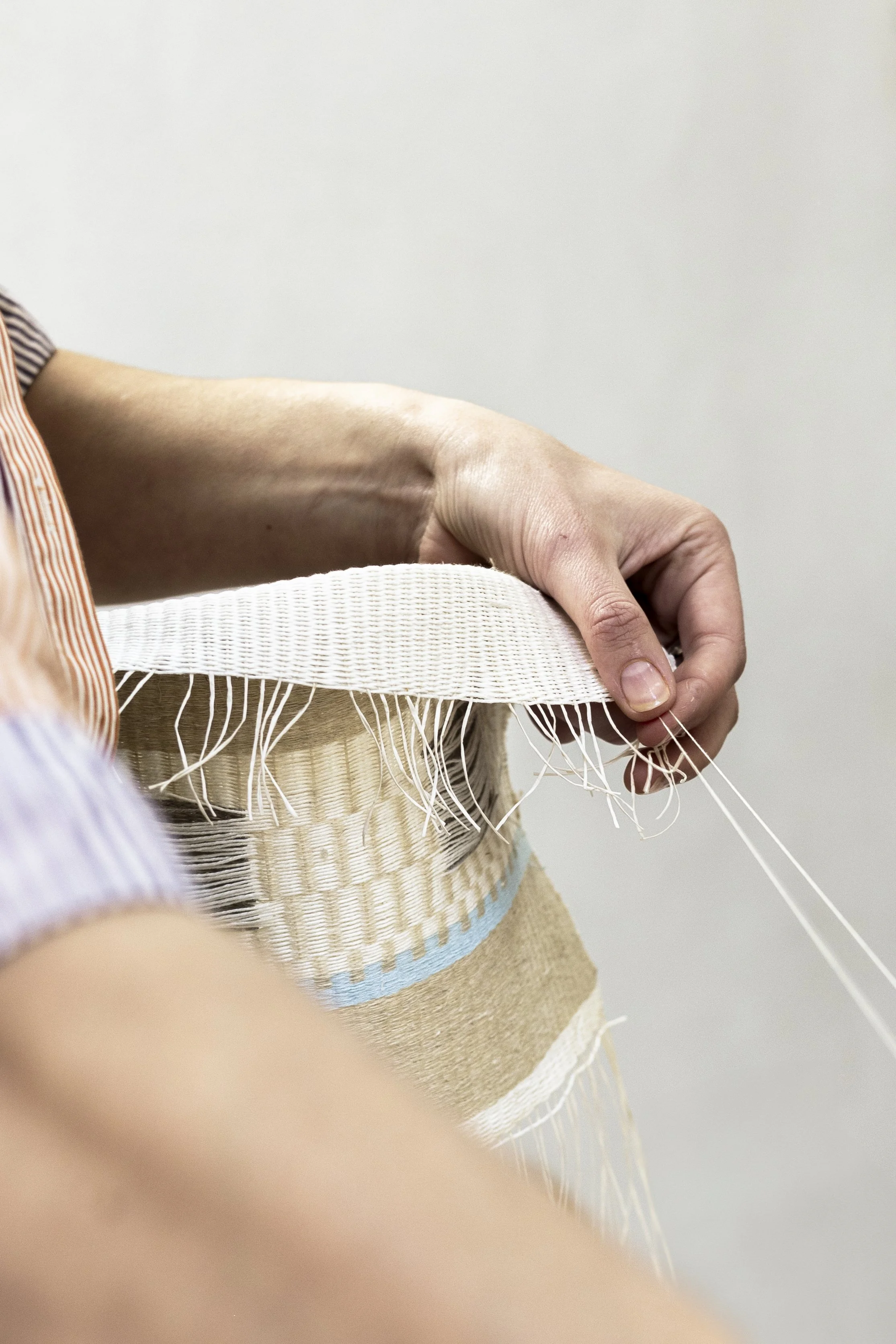 Une main qui tire sur un fil pour un travail de tissage ou de fabrication artisanale, avec un vêtement rayé à manches courtes visible.