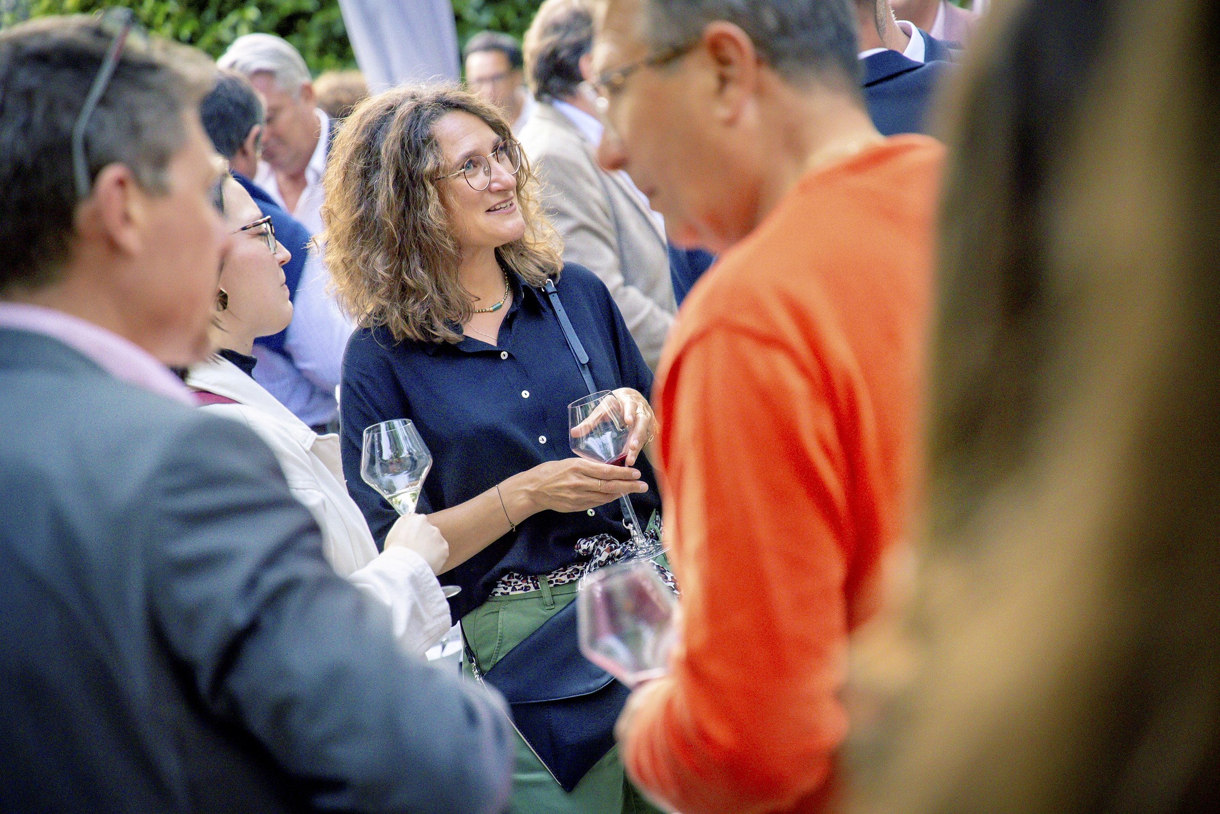 Une femme aux cheveux bouclés, portant des lunettes et un chemisier bleu, tient un verre de vin rouge lors d'un événement social en plein air, entourée de plusieurs personnes.