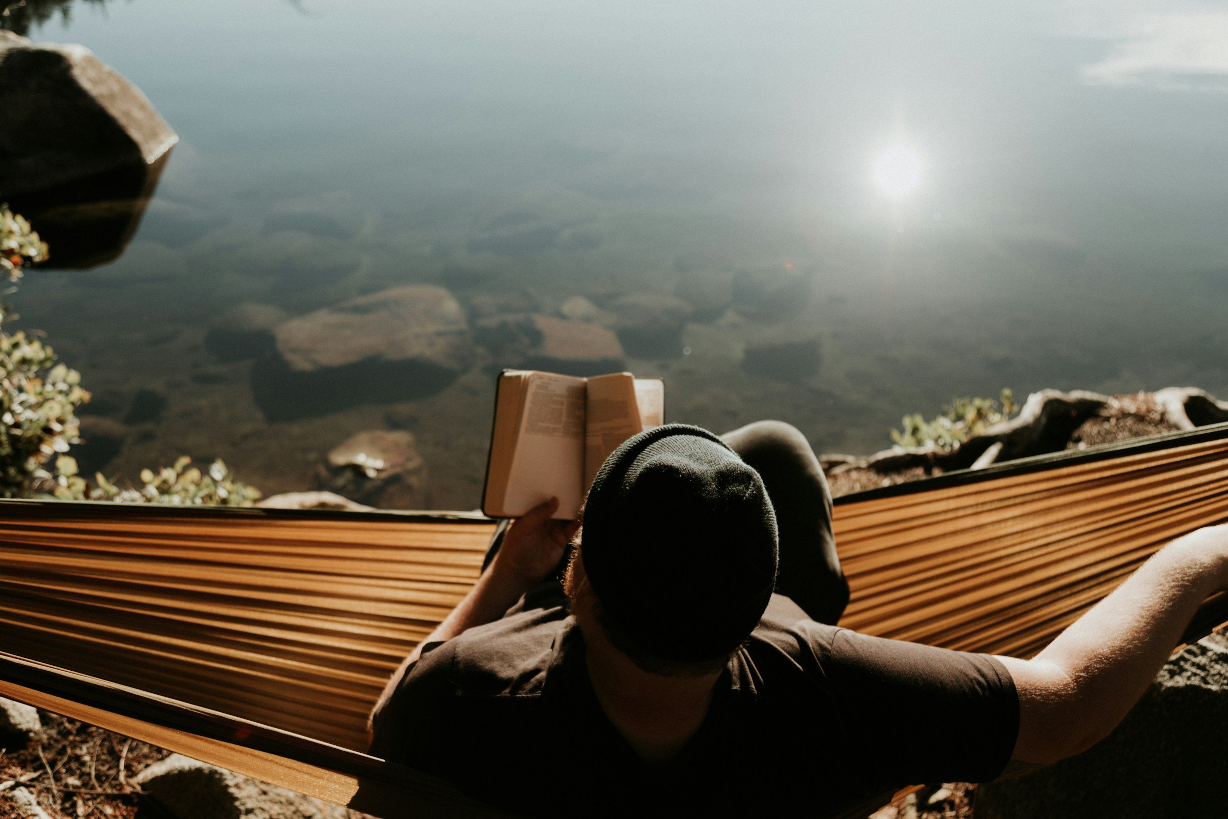 Person sitting in a hammock, reading a book, beside a lake or river with rocks and plants visible in the water. Therapy with me is collaborative, warm, and grounded in real-life application.