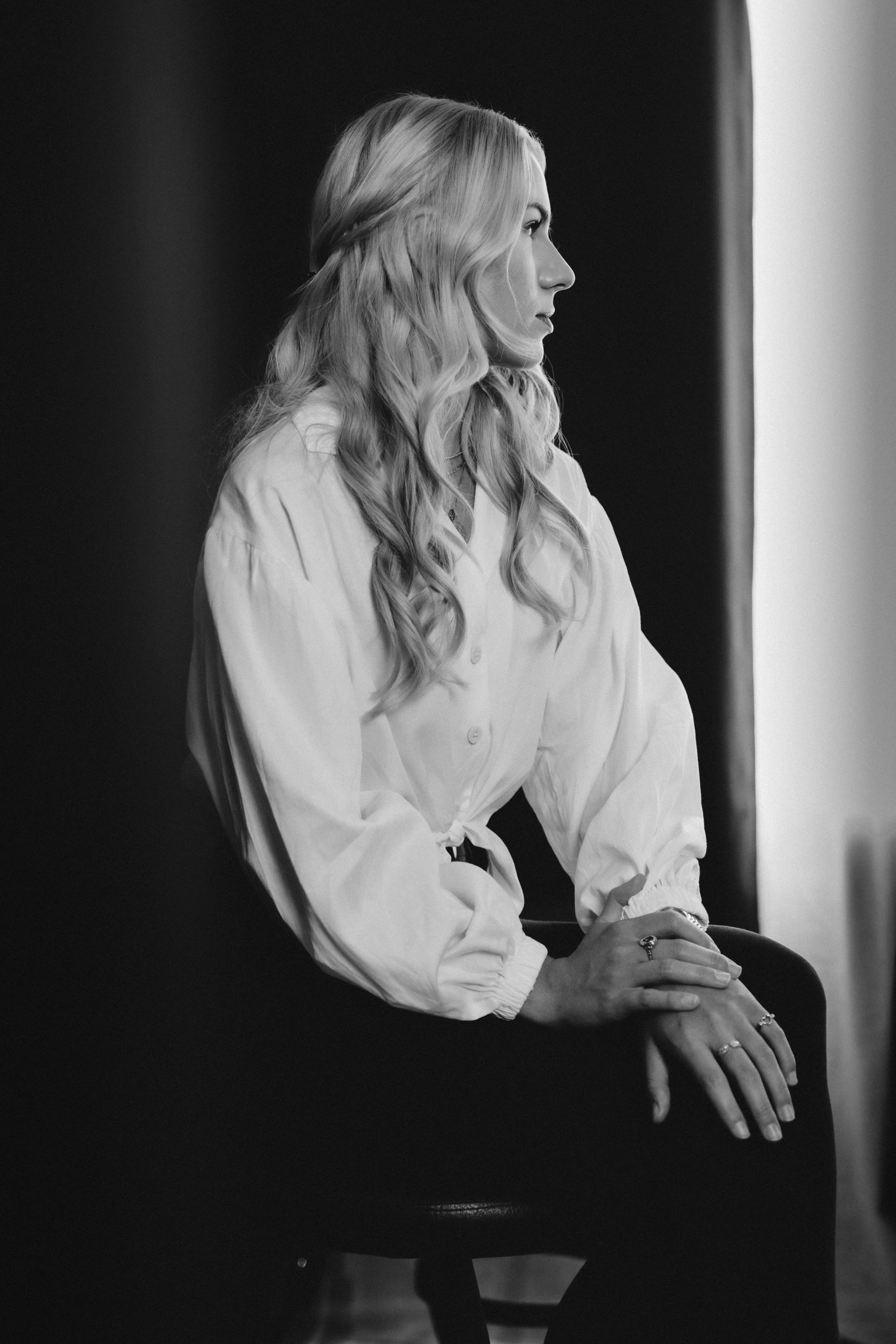 A black-and-white photo of a woman with long, wavy hair sitting on a chair, looking to her right, wearing a white blouse with puffy sleeves, with her hands resting on the chair, and appears to be in a contemplative mood.