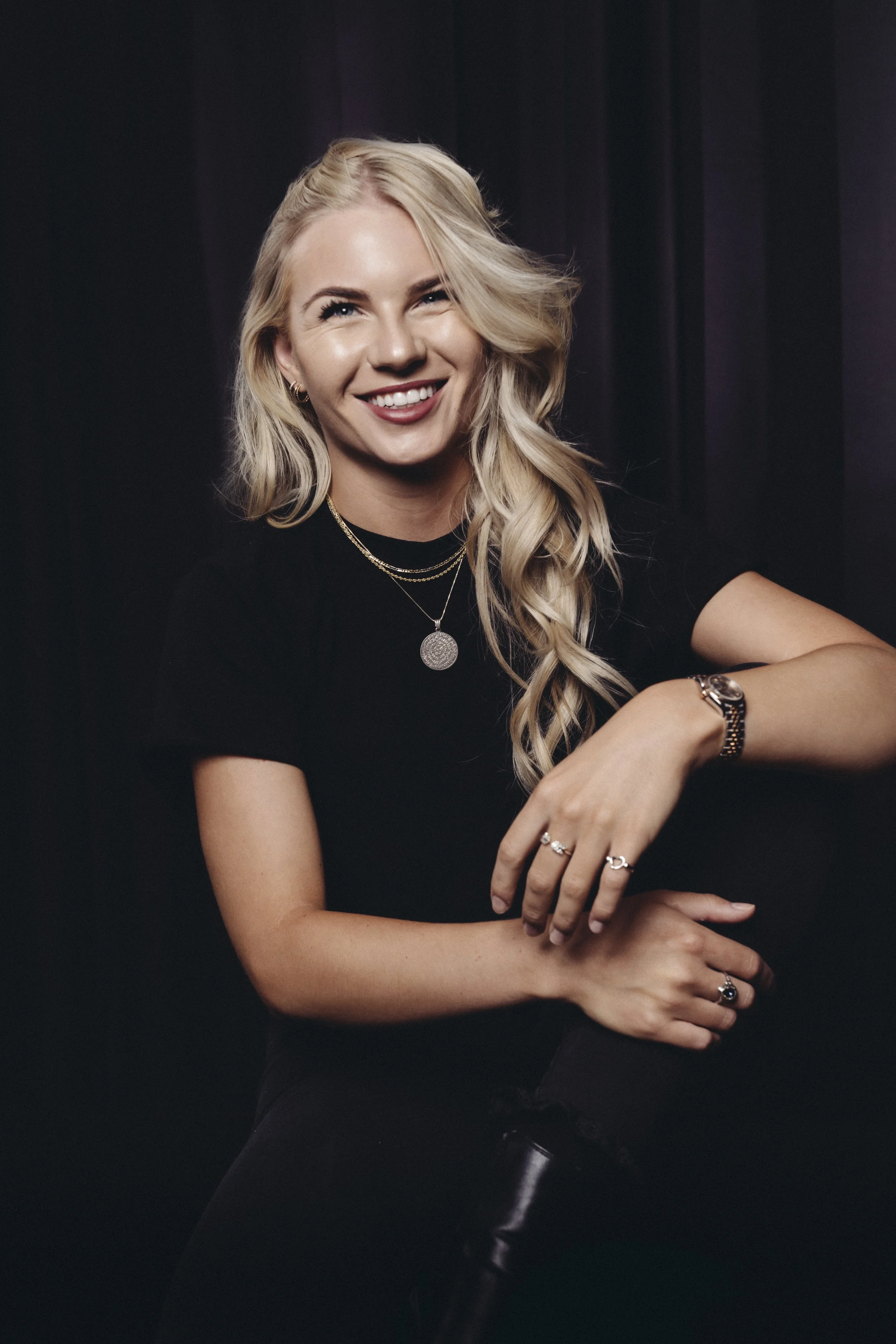 A young woman with long, wavy blonde hair, smiling while wearing a black top and jewelry, against a dark background.