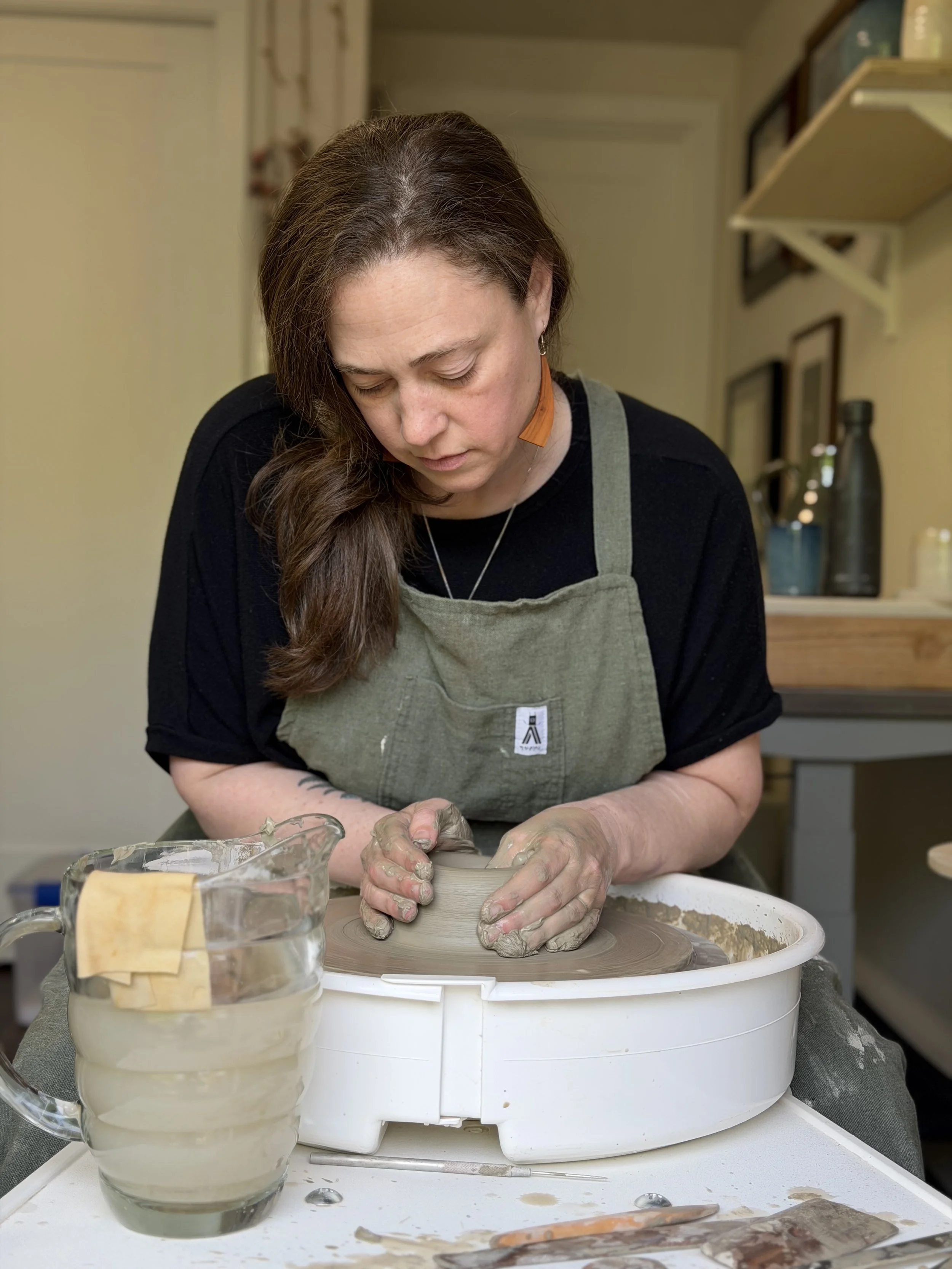 A woman with brown hair and earrings is working on a pottery wheel, shaping clay with her hands in a ceramic studio. There are cups of water and tools on the table in front of her.