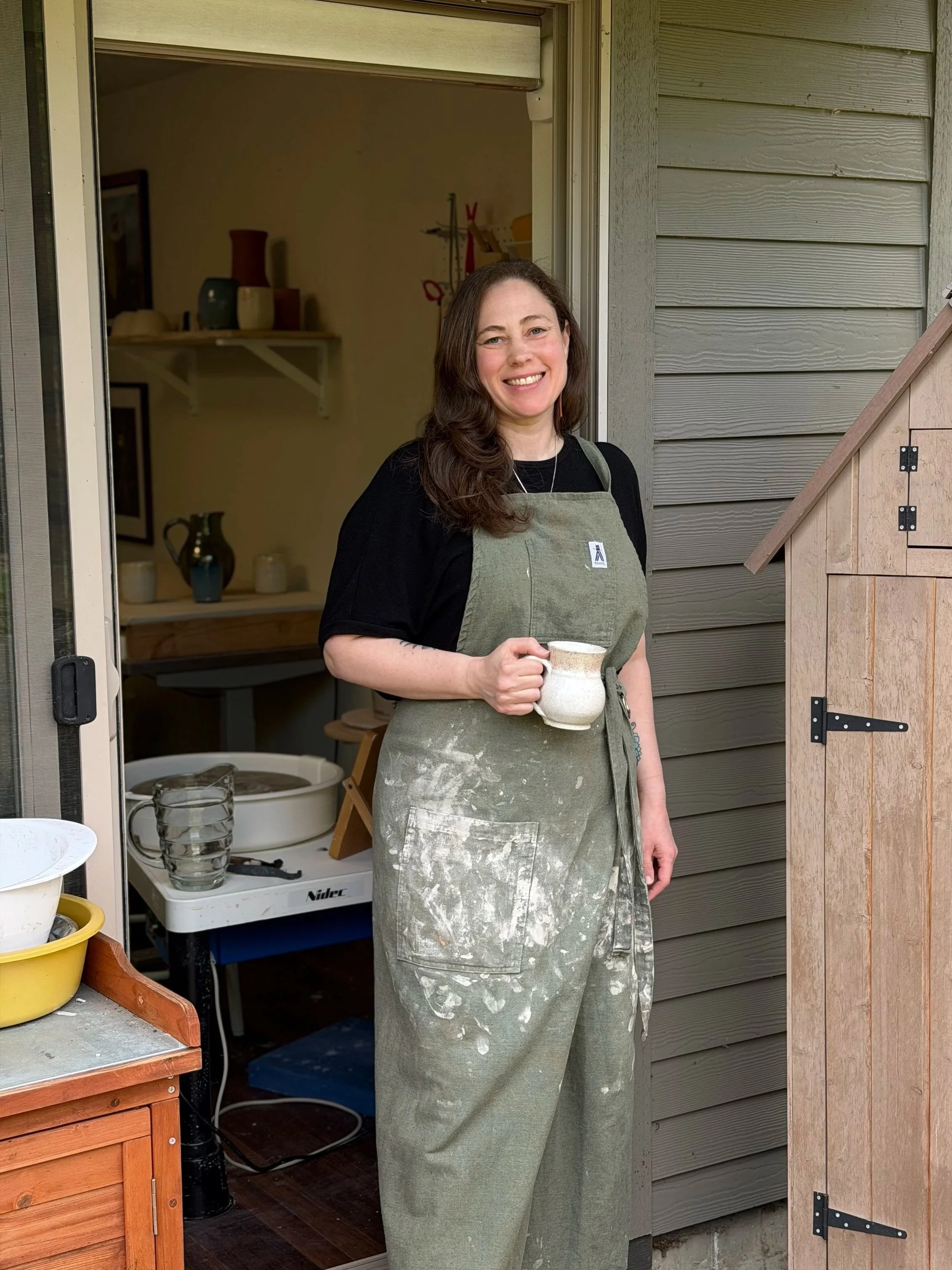Woman smiling in doorway wearing a black shirt and an apron holding a ceramic mug, with a wooden shed and ceramic items on a table behind her.