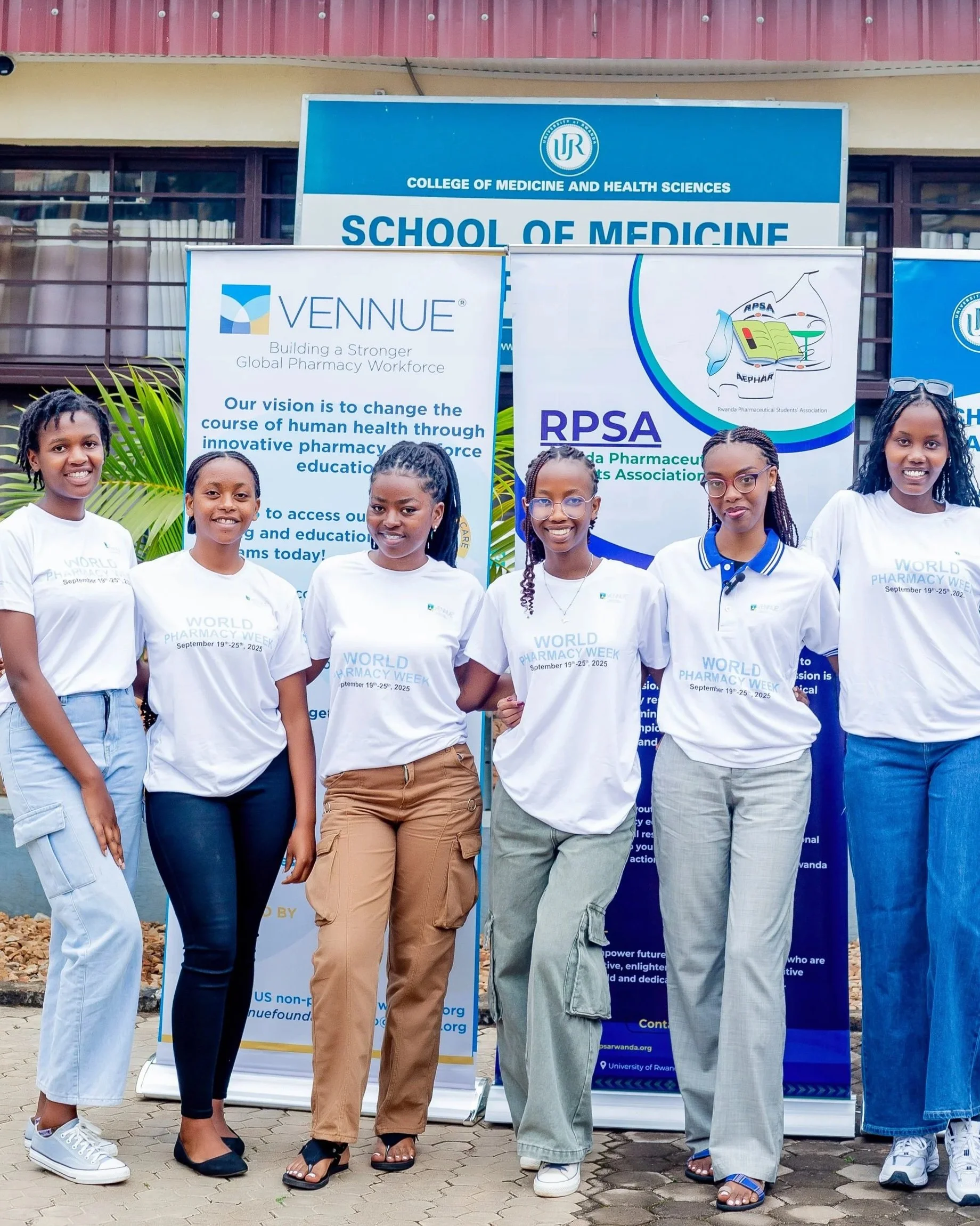 Six young women standing in front of banners at a university event celebrating World Pharmacy Week, September 19-25, 2025, wearing white t-shirts with event details.