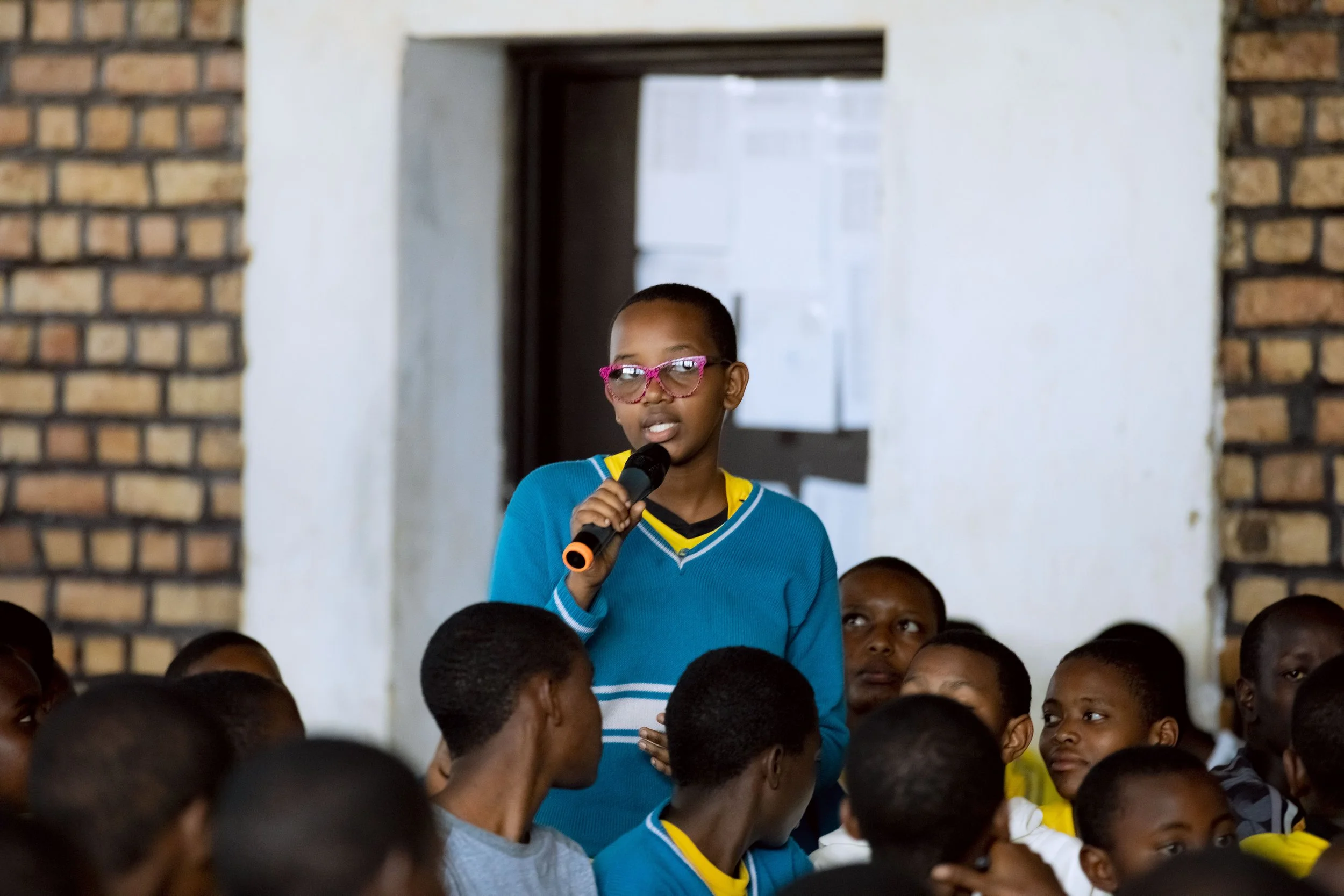 A young girl wearing pink glasses and a blue sweater is standing and speaking into a microphone in a room with many seated children, a mixture of boys and girls, some looking at her and others looking away, with a background of a white and brick wall.
