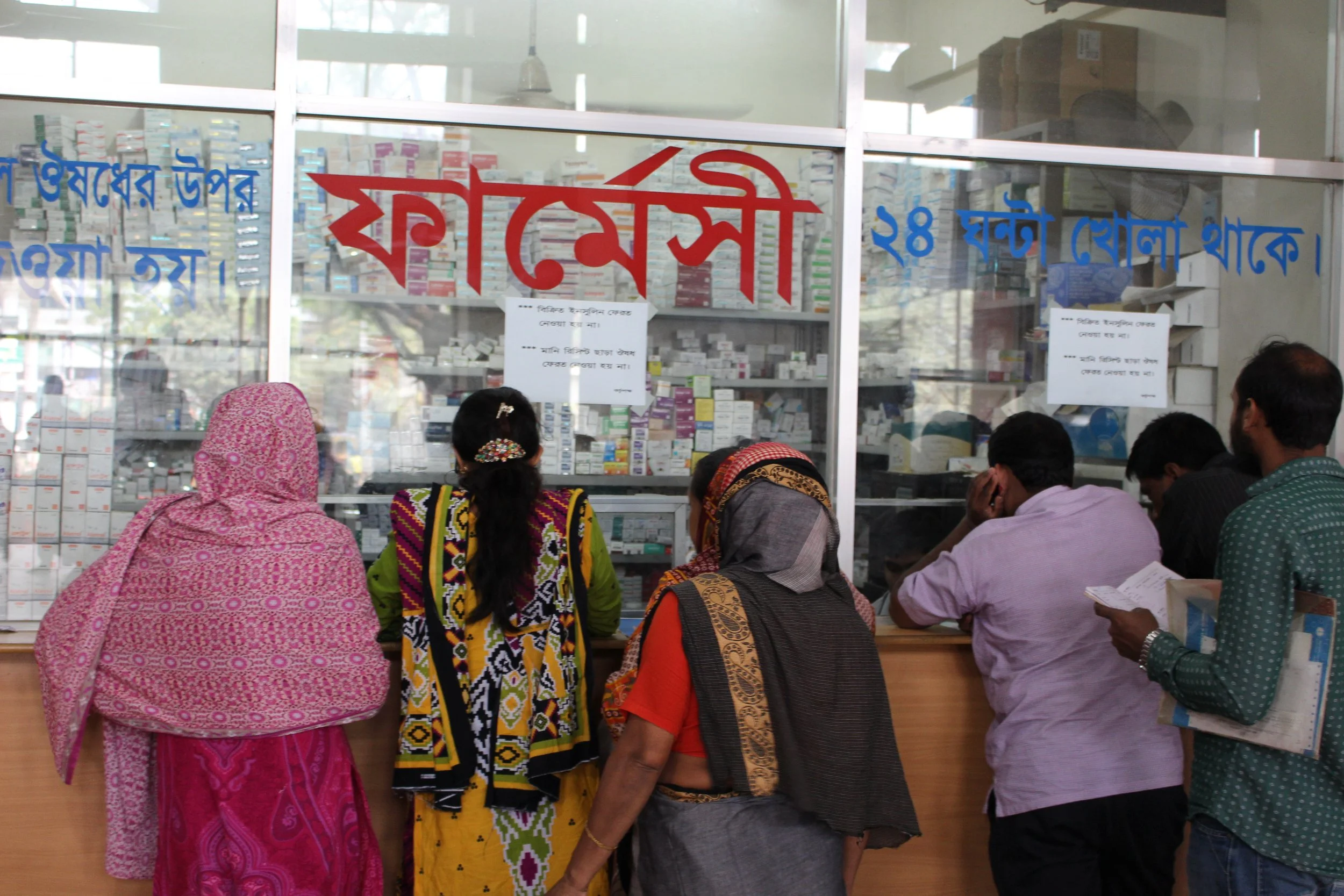 People standing at a pharmacy counter inside a store, with shelves stocked with medicines visible through the glass behind the counter.