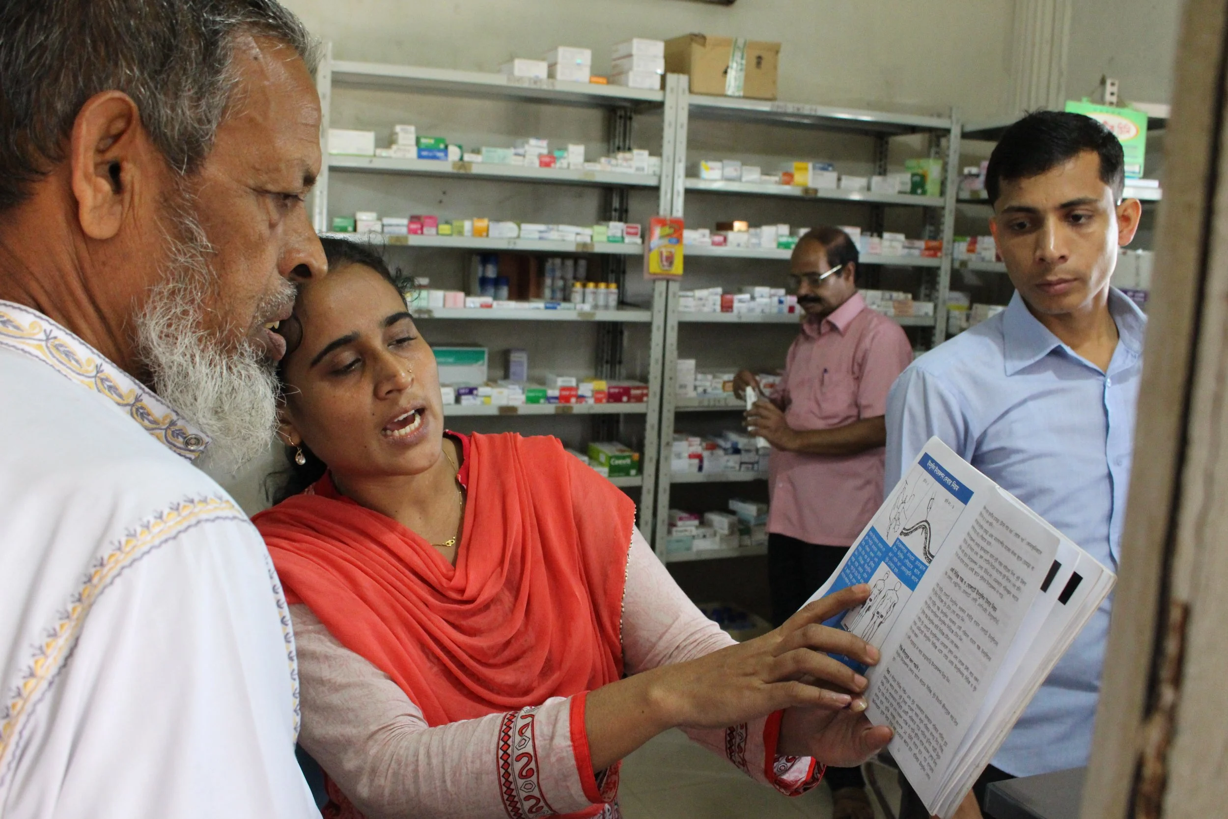 A woman in a red saree showing prescription information to an elderly man with a white beard in a pharmacy. A young man in a light blue shirt observes while a man in a pink shirt looks at some medication in the background.