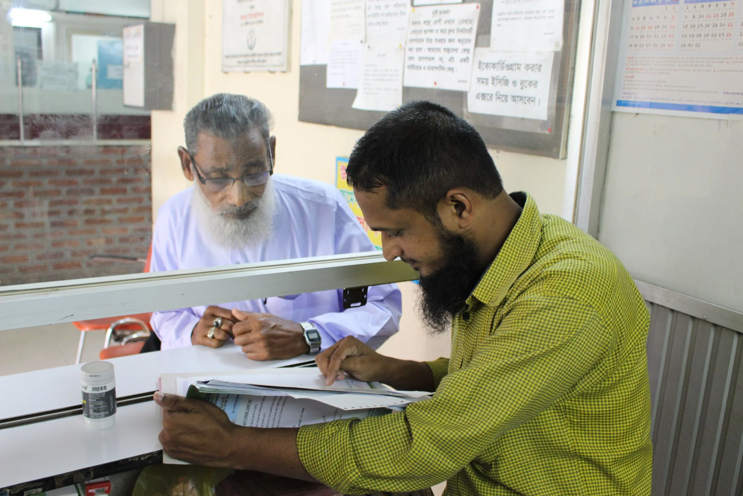 An elderly man with a white beard standing behind a pharmacy partition. A younger man with a beard and wearing a yellow checked shirt is sitting at a desk, reviewing documents. There are posters and notices on the wall behind them.