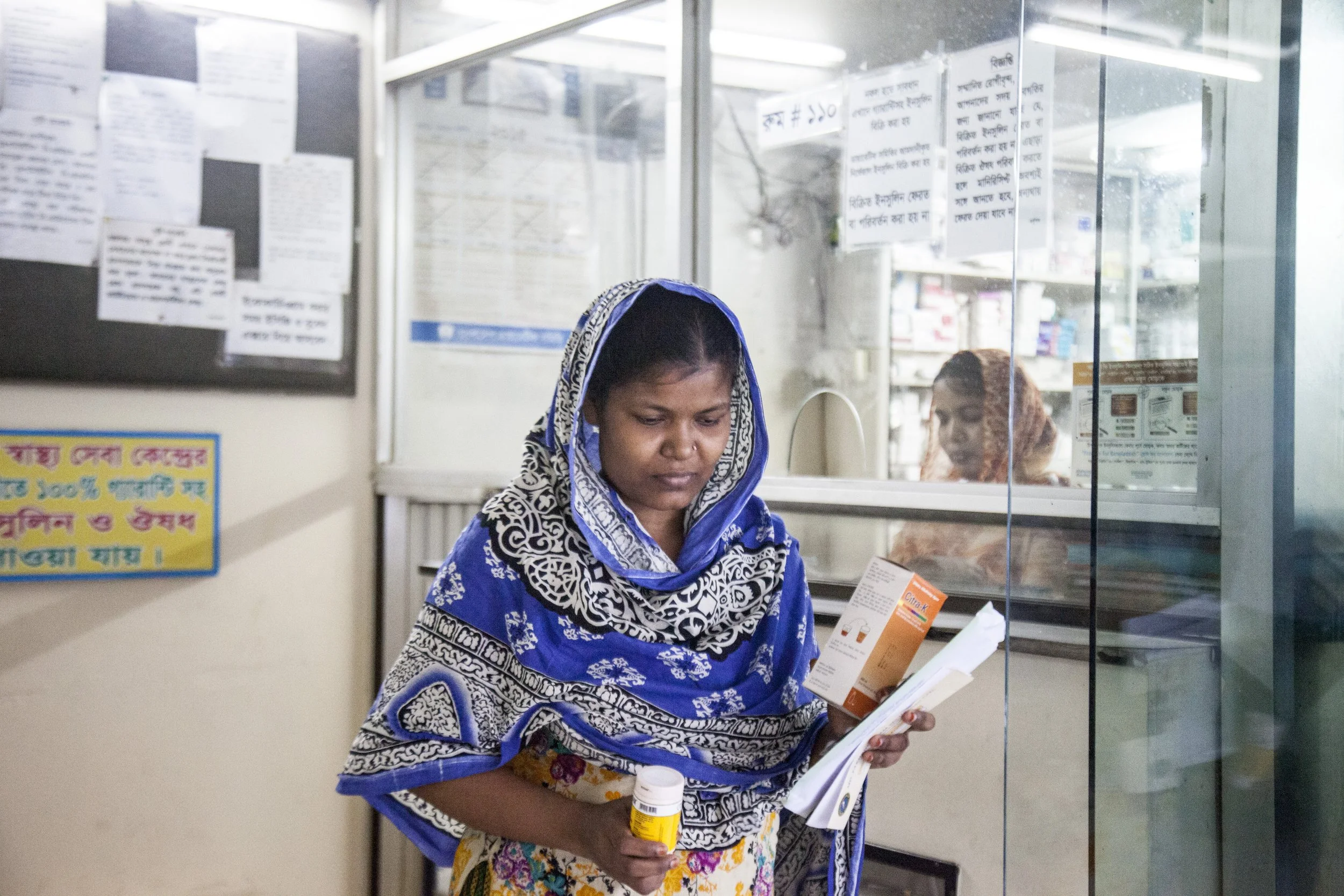 A woman wearing a blue patterned headscarf stands in a pharmacy, looking at a pamphlet and holding a yellow medication bottle. In the background, another woman is visible through the glass window of the pharmacy.