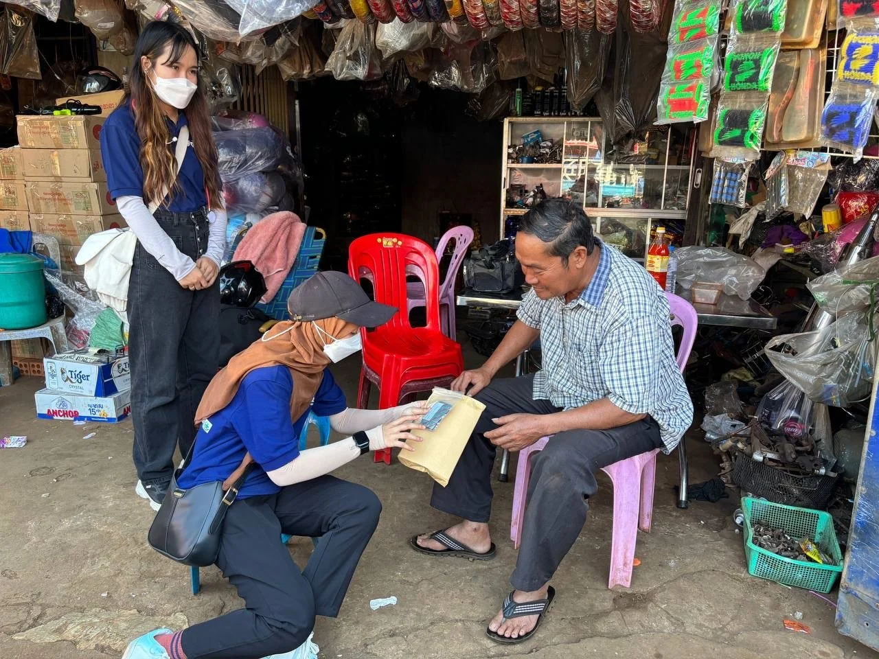 A woman in a blue shirt handing literatur to a man sitting on a pink chair in a rural village. Another woman, stands nearby observing. The market stall behind them is filled with various goods, including plastic bags, boxes, and tools.