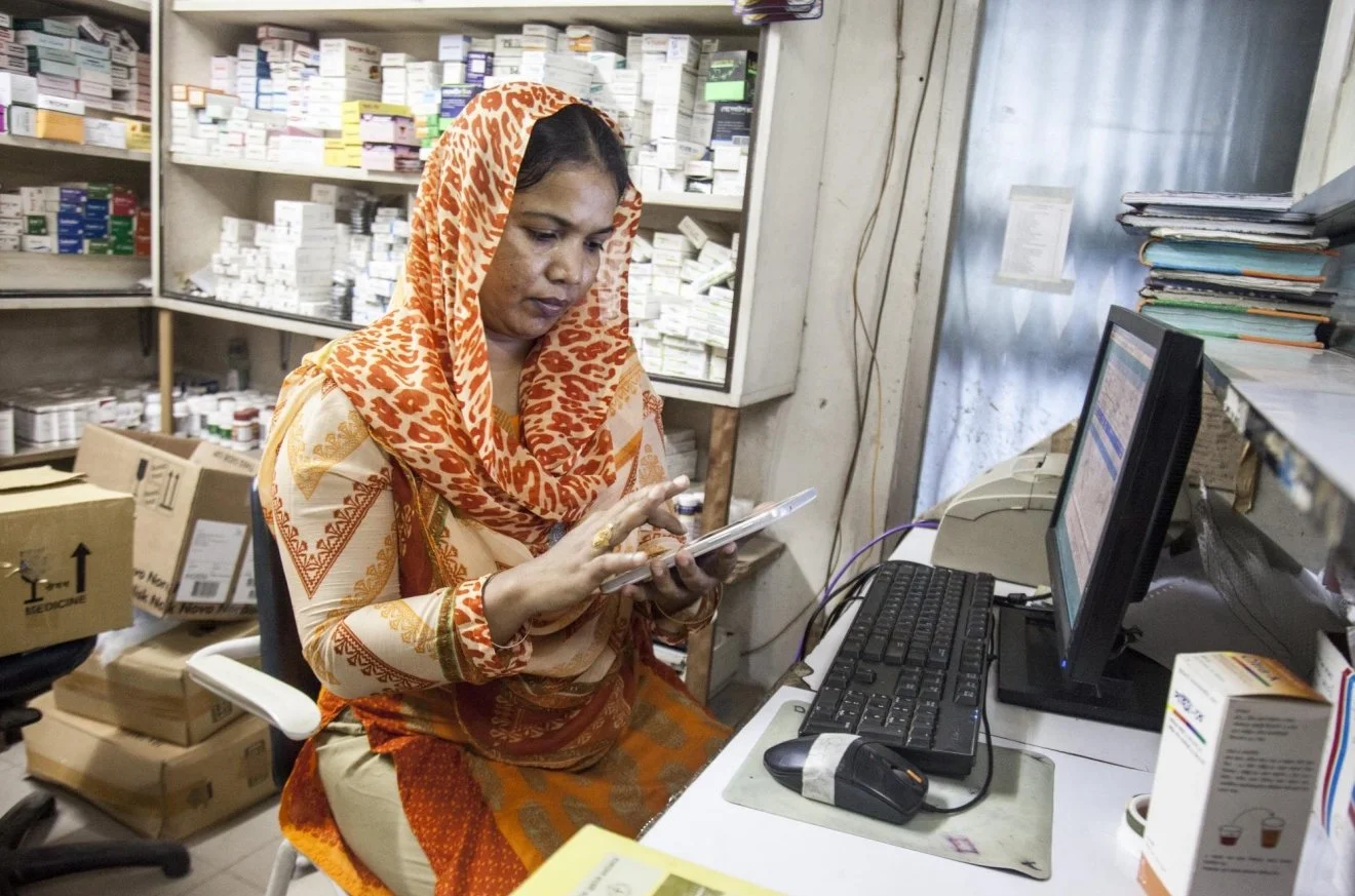 A pharmacist in a patterned orange and cream traditional attire sitting at a cluttered desk, looking at her phone, with shelves of medicine bottles and boxes behind her.