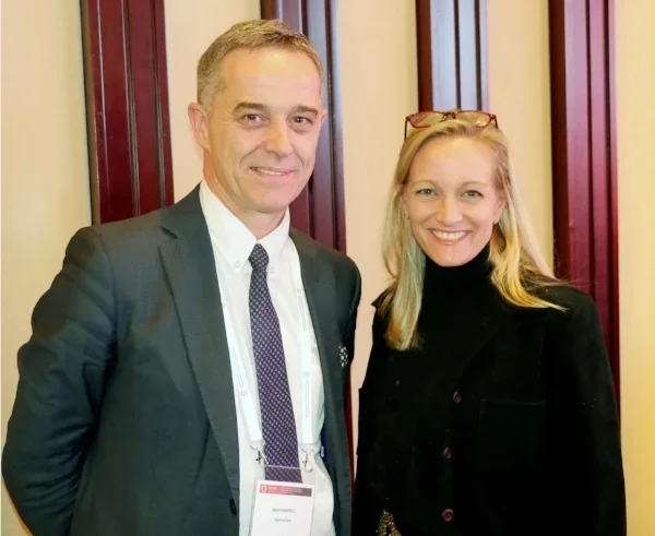 Vennue's Tammy Allen with the WHO's Jim Campbell smiling and posing together indoors. Campbell is wearing a suit and tie, and the Tammy is in a black top and jacket with glasses resting on her head.