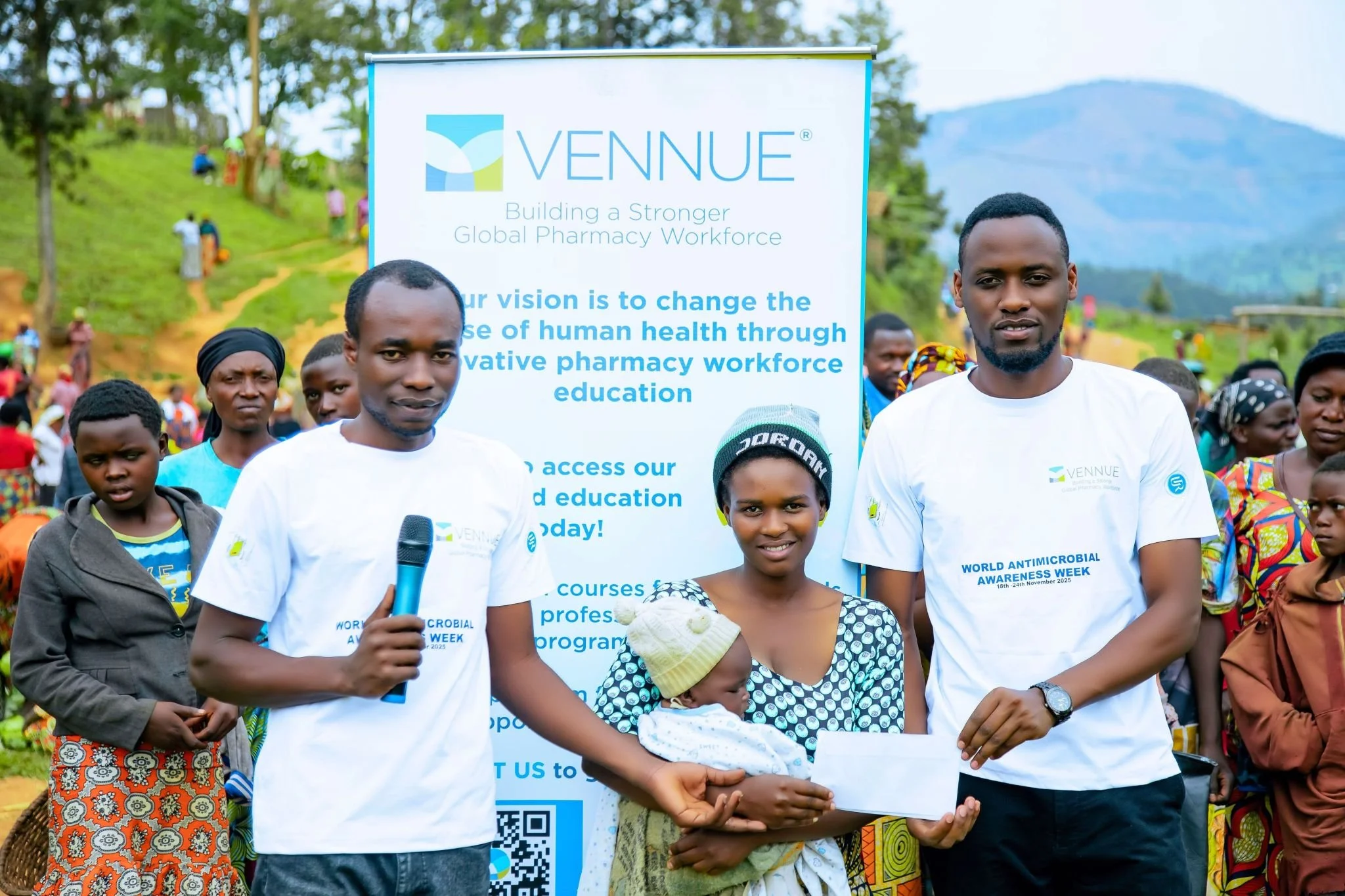 A group of people gathered outdoors in front of a banner that reads 'Vennue, Building a Stronger Global Pharmacy Workforce' and promotes education. Two men in white Vennue t-shirts are standing on either side of a woman holding a baby, with one man holding a microphone and the other holding an envelope. There are children and women in colorful clothing in the background, with a hilly landscape and trees.