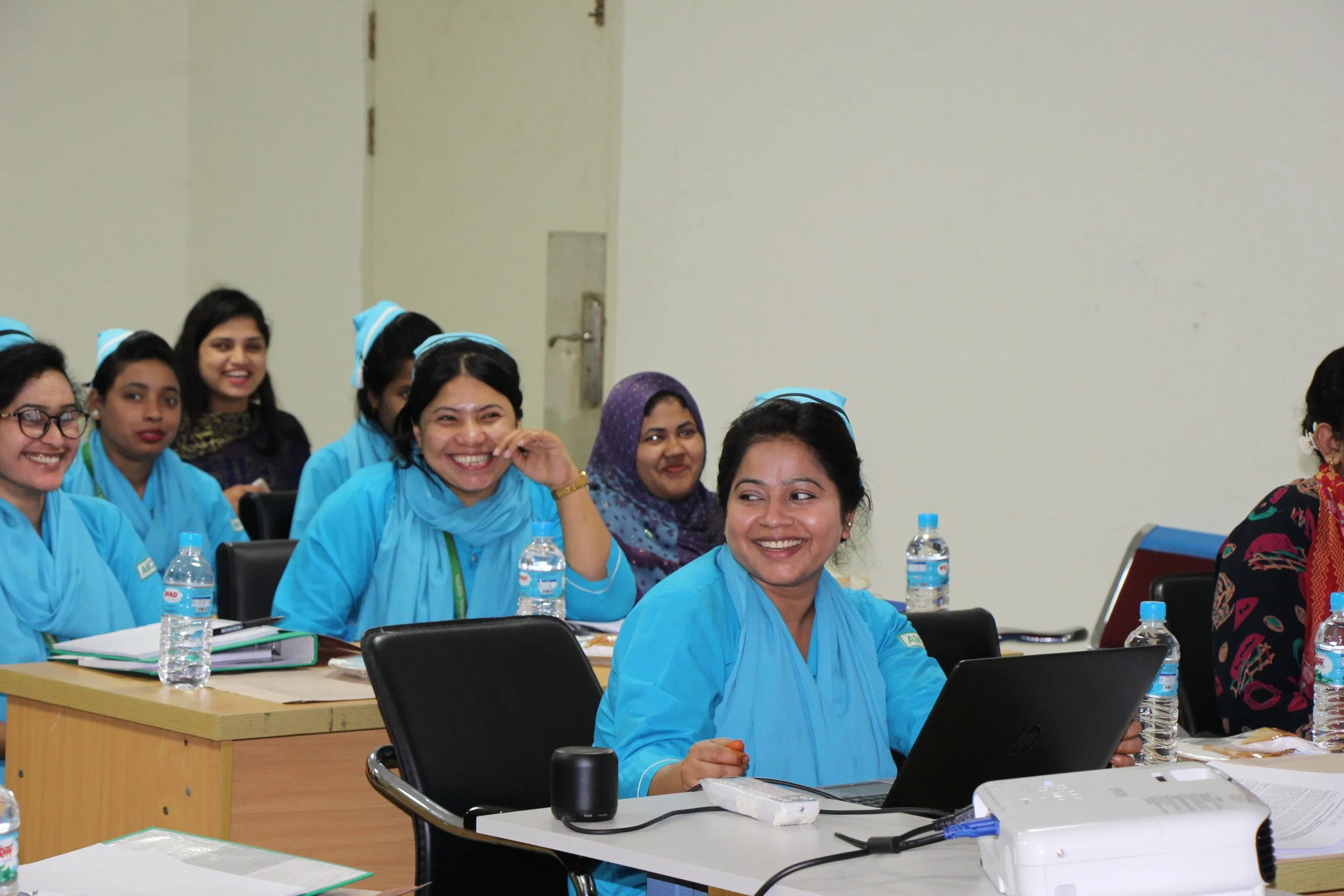 Group of women in blue uniforms attending a meeting or training session, smiling and engaging with each other, with notebooks, water bottles, and a laptop on the table.