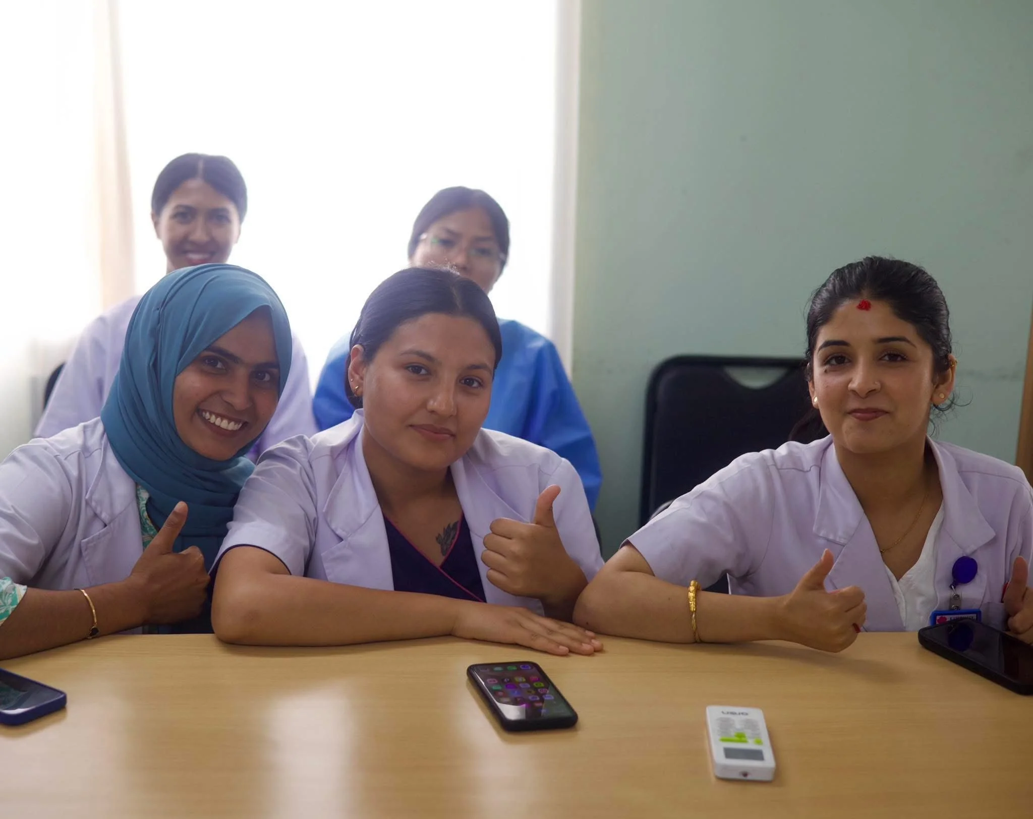Group of five women in medical uniforms sitting at a table indoors, smiling and showing thumbs up, with mobile phones on the table.