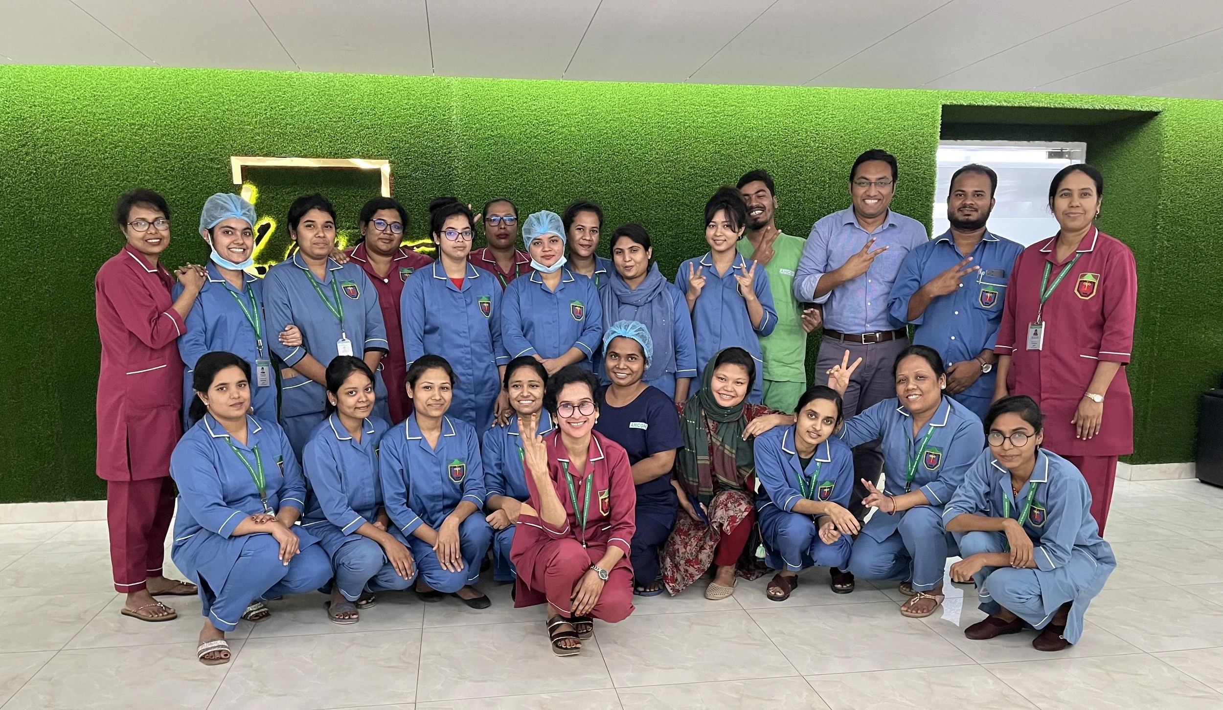 A group of healthcare professionals and staff, including men and women of diverse ethnicities, pose together indoors in front of a green wall with a logo. They are dressed in scrubs, some in blue and others in maroon, with some wearing hairnets and masks, and others wearing ID badges, smiling and making hand gestures, indicating a team or staff photo.