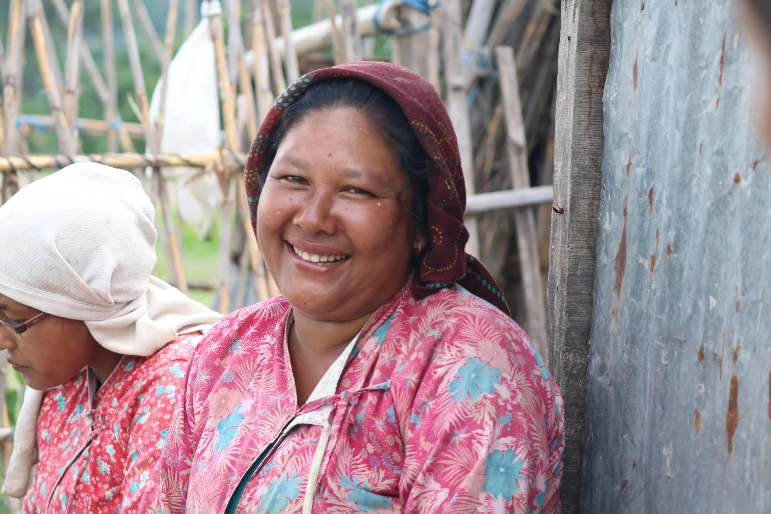 Smiling woman in pink floral shirt with headscarf standing outdoors near wooden structure, with another person in a white head covering nearby.