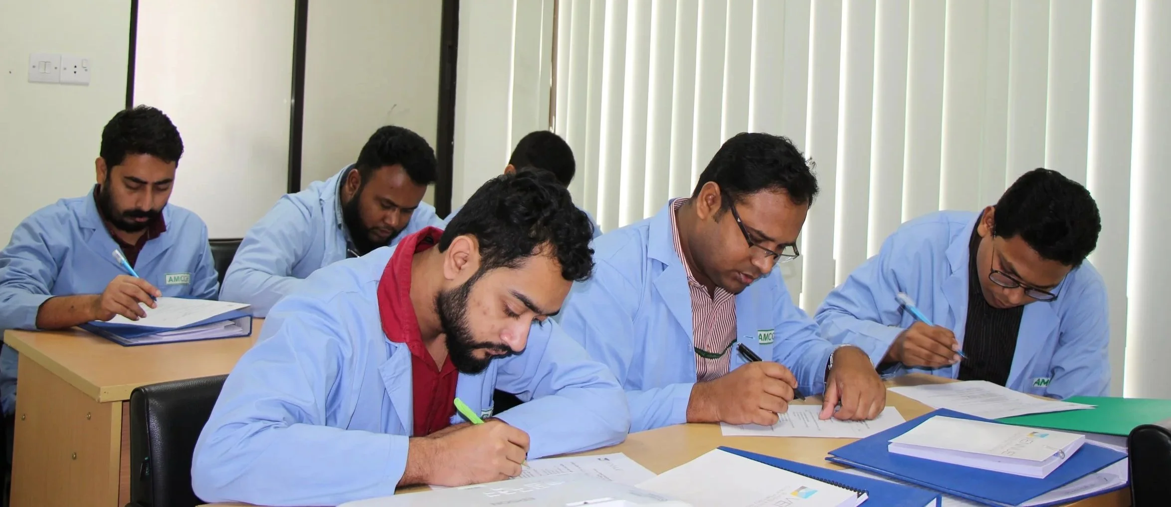 Group of pharmacist trainees in light blue lab coats sitting at desks and taking notes during training or lecture in a classroom.