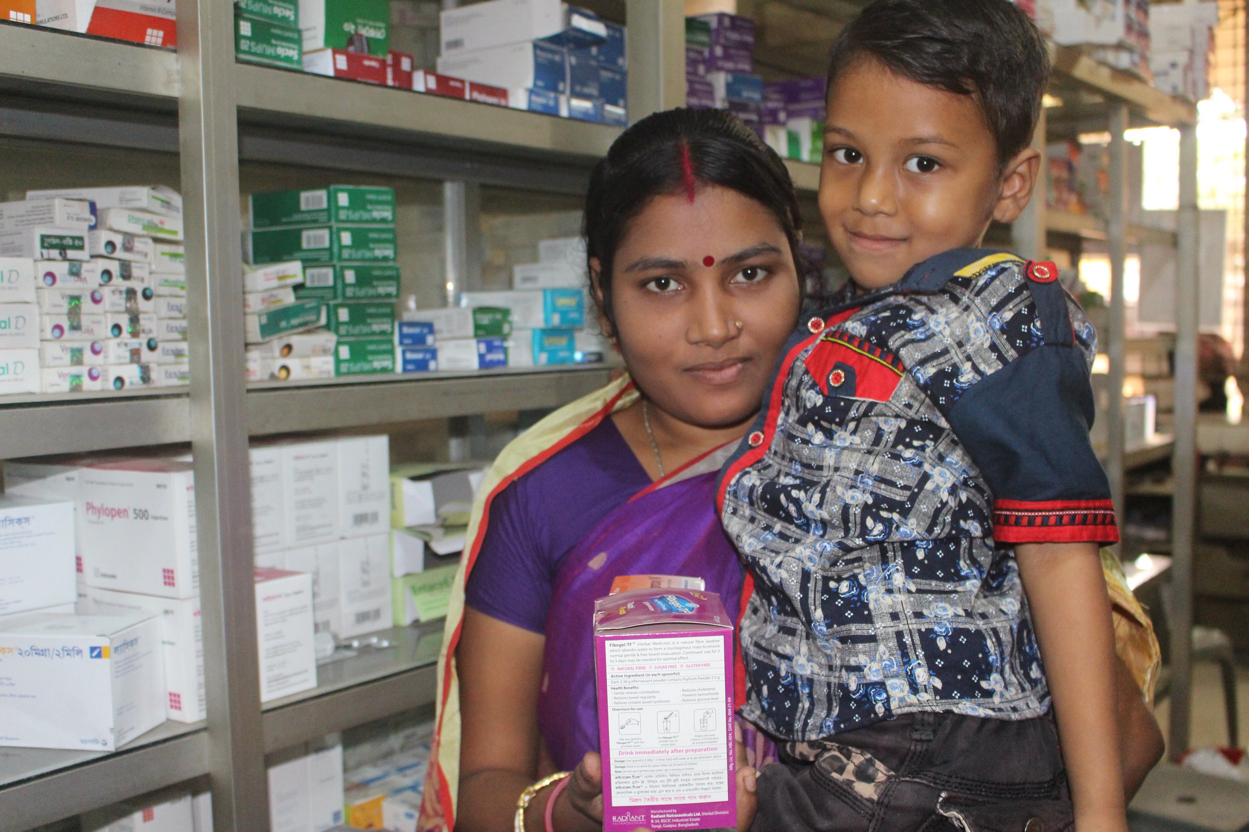 A woman in a purple saree holding a child in a patterned shirt and shorts in a pharmacy with shelves of medicines behind them.