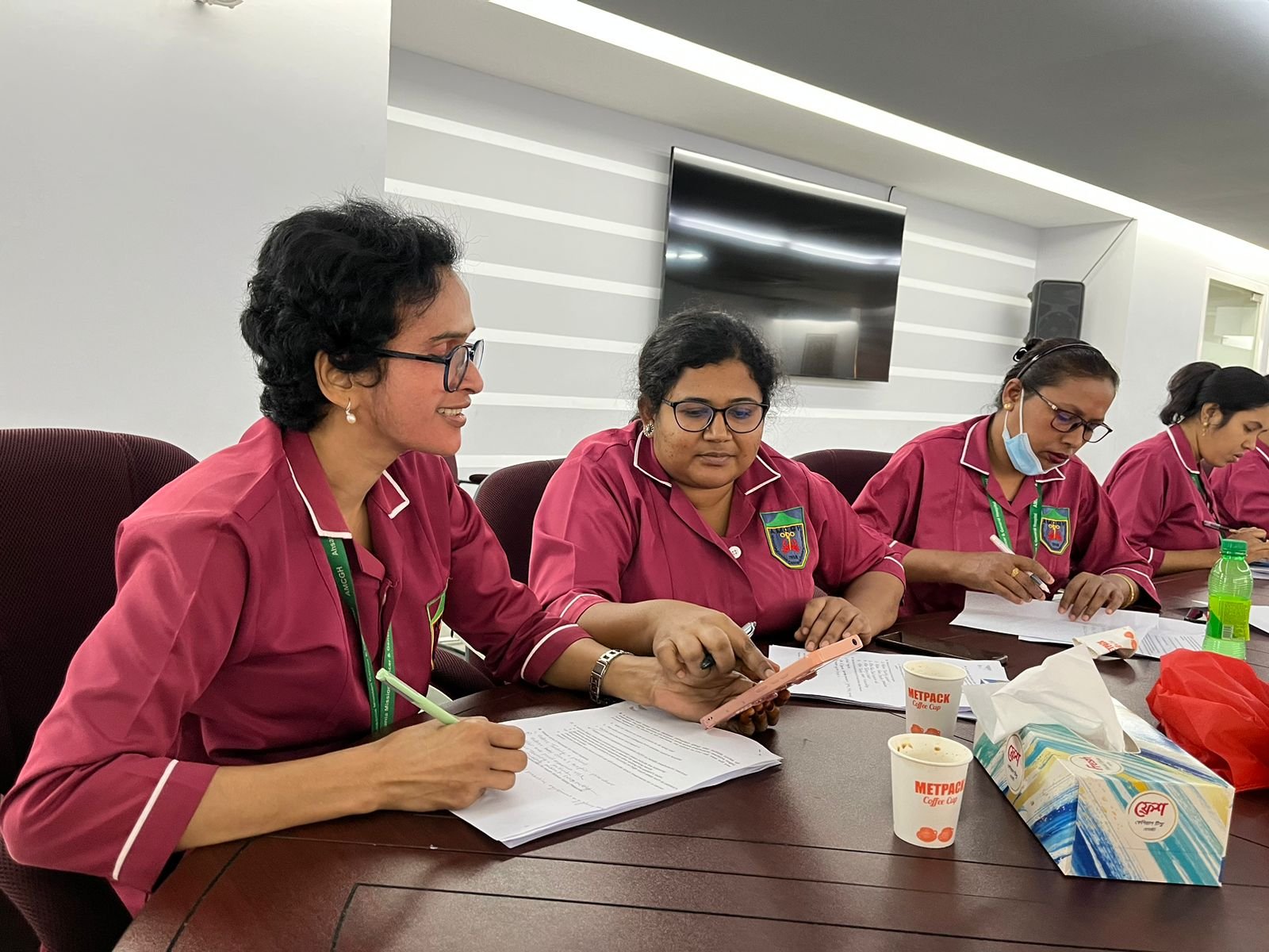 Four cancer-care nurses in pink uniforms sitting at a conference table, reviewing documents and using a phone, with tissue box and cups on the table in a modern conference room.