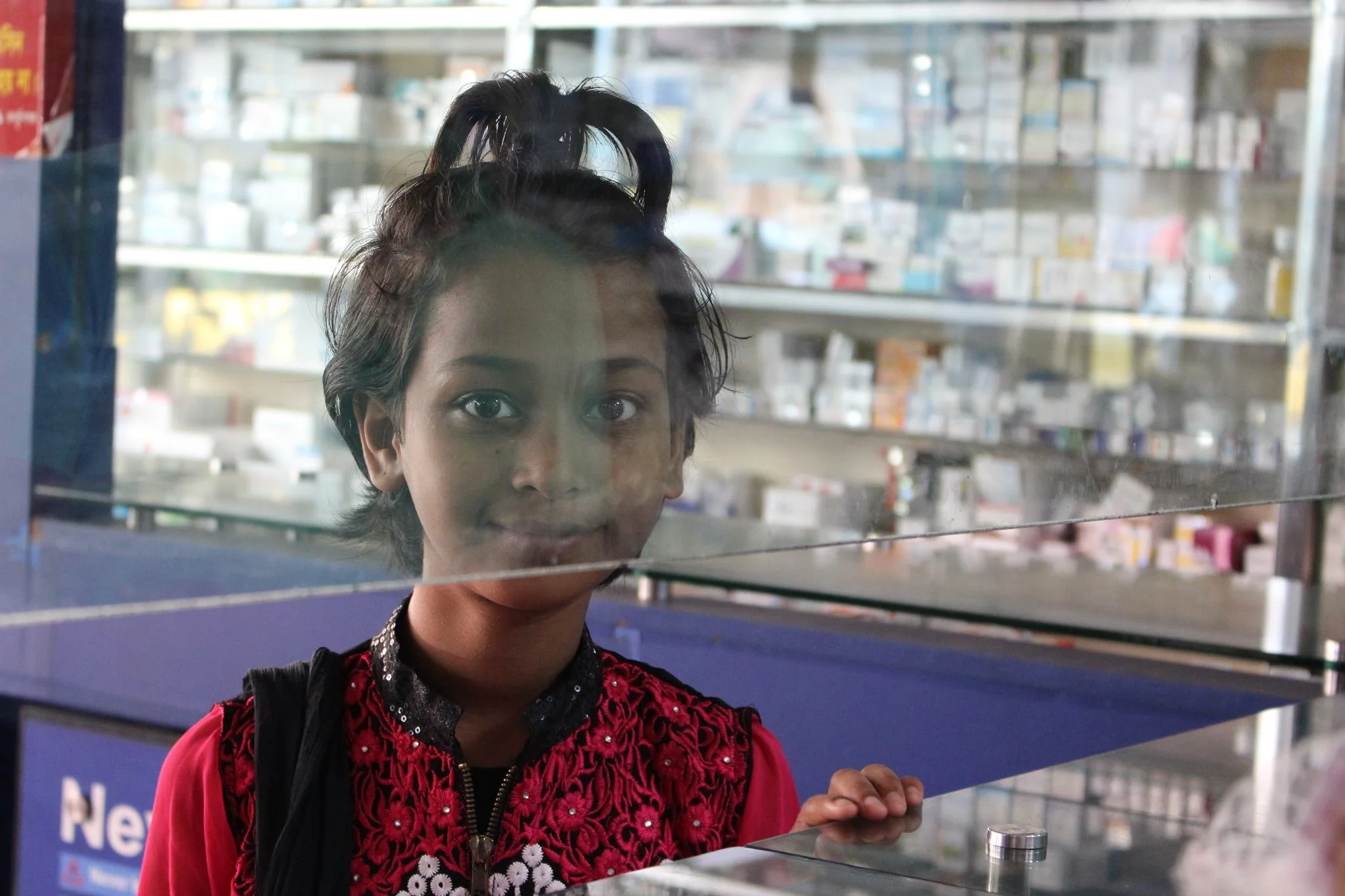 A young woman with short dark hair and a red embroidered jacket gazes through a pharmacy window. She is standing in front of shelves filled with medicines and pharmacy supplies.