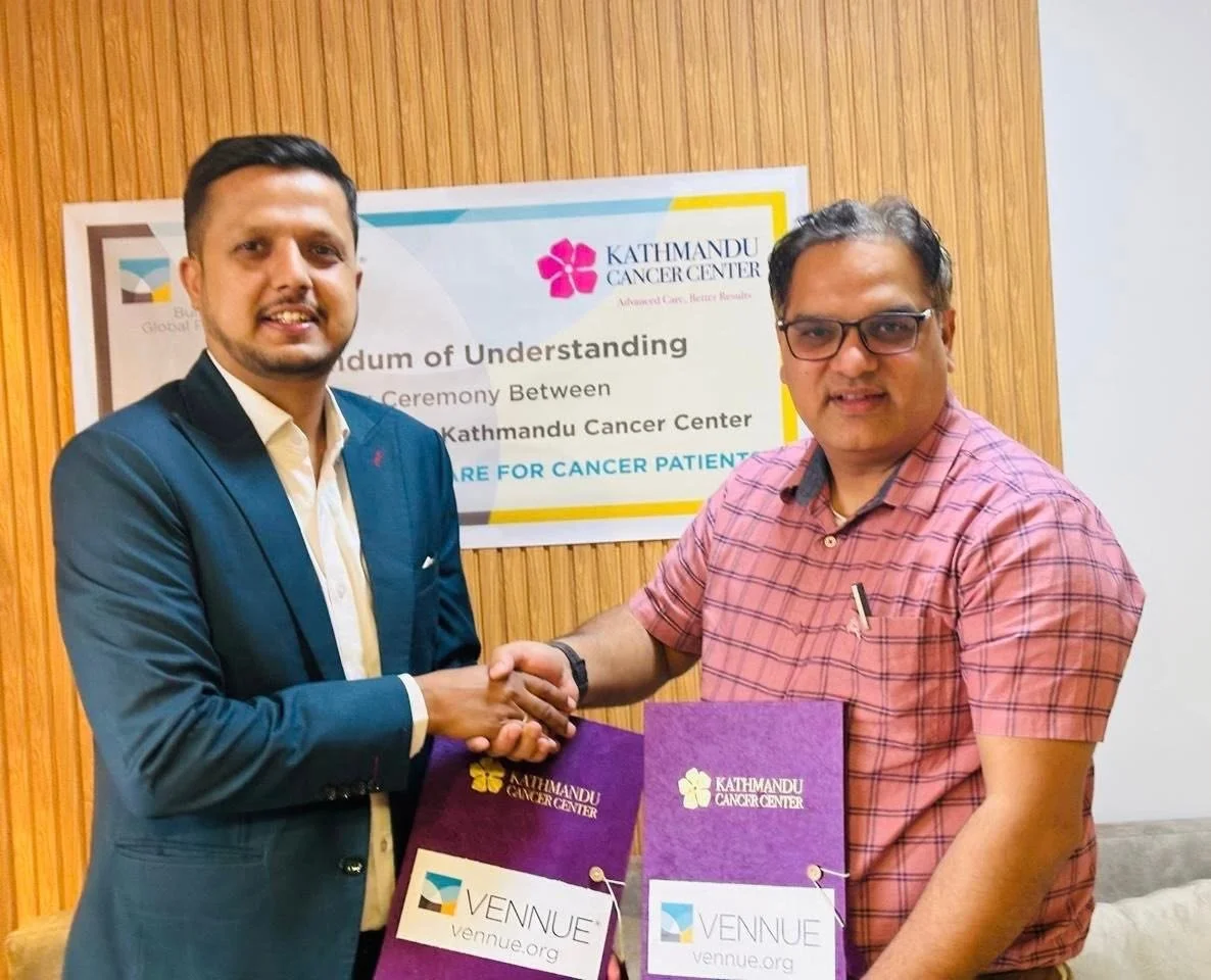 Two men shaking hands in front of a sign at Kathmandu Cancer Center, holding purple folders with the center's logo, smiling, in a room with wooden wall paneling.