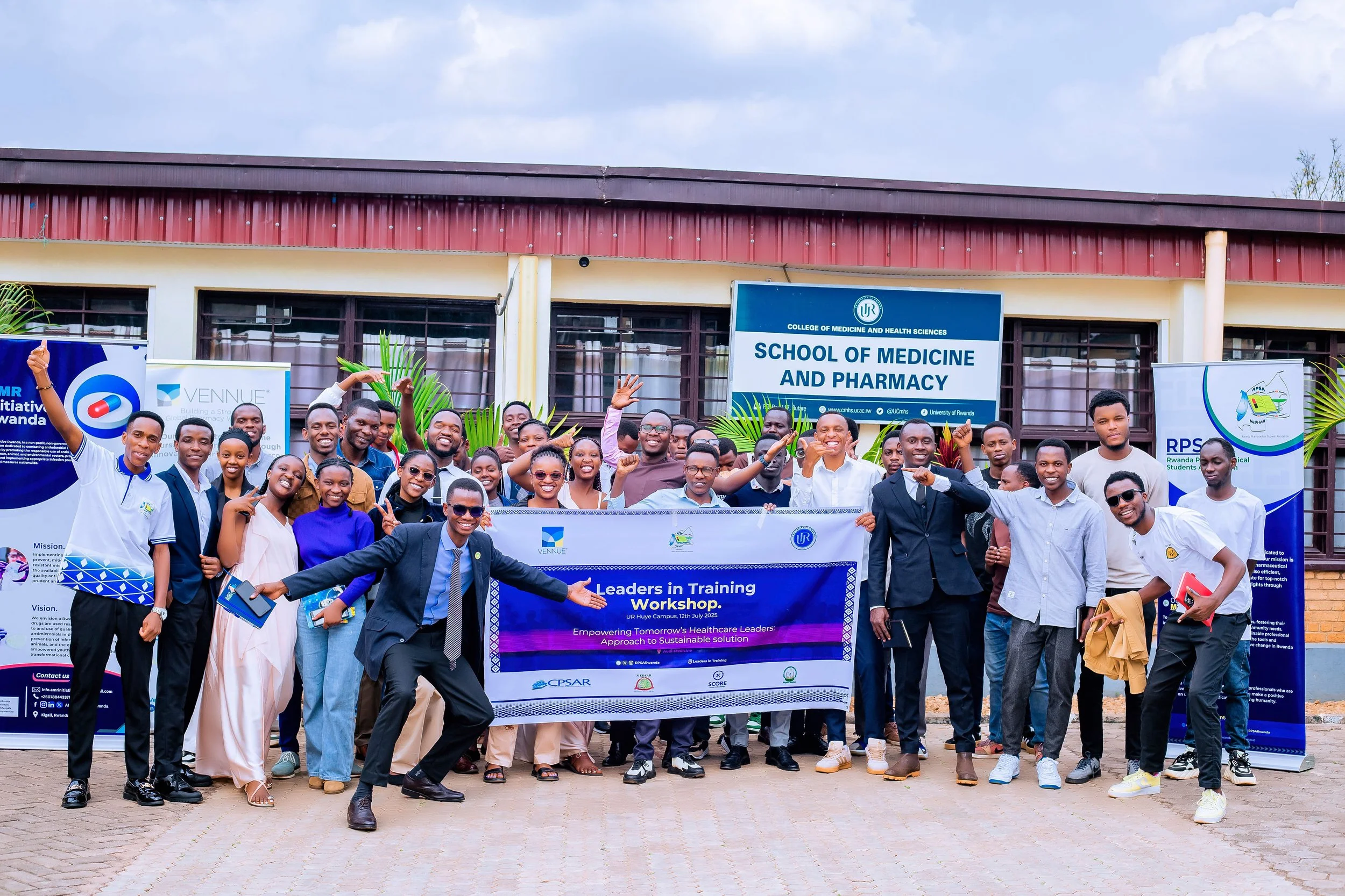 Group of people standing outdoors in front of a building with a sign that reads 'School of Medicine and Pharmacy,' holding a banner for a training workshop. The group is smiling, some making peace signs, dressed in formal and casual clothing.