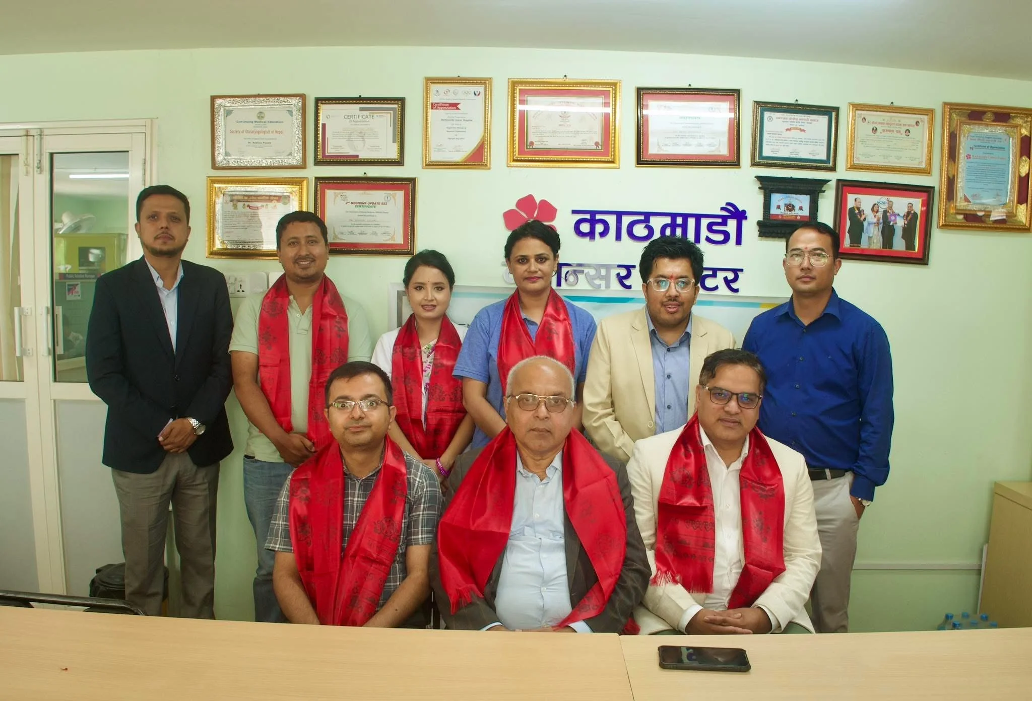Group of nine people, six men and three women, posing for a photograph in an office with framed certificates on the wall and a sign with Nepali text behind them. Some individuals are wearing red scarves.