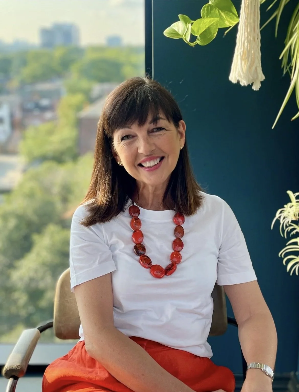 This is Tracy Smithers, Executive Coach. A smiling woman with dark brown hair, wearing a white t-shirt and a red necklace, sitting indoors near a large window with a cityscape and green trees in the background, and some hanging plants visible.