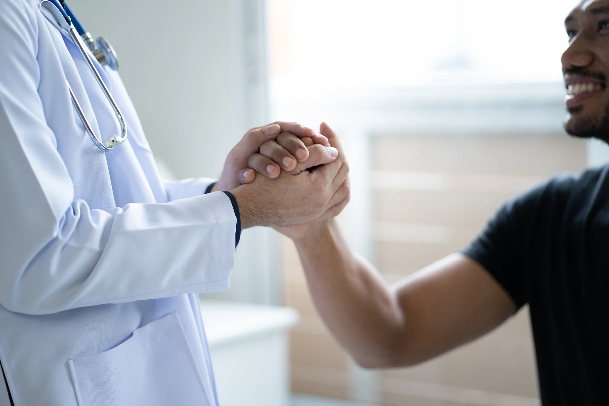 A doctor in a white coat shaking hands with a smiling patient in a black shirt in a bright medical office.