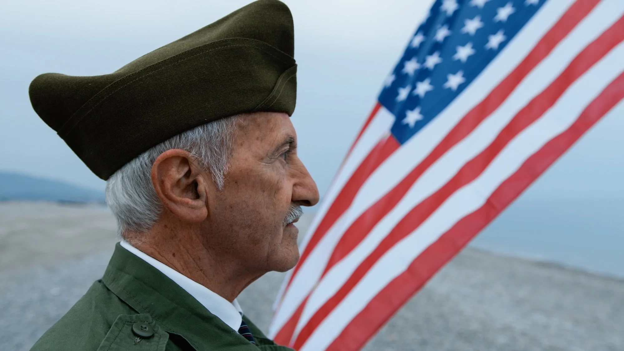 An elderly man in military uniform holding an American flag on a beach, looking solemnly at the flag.
