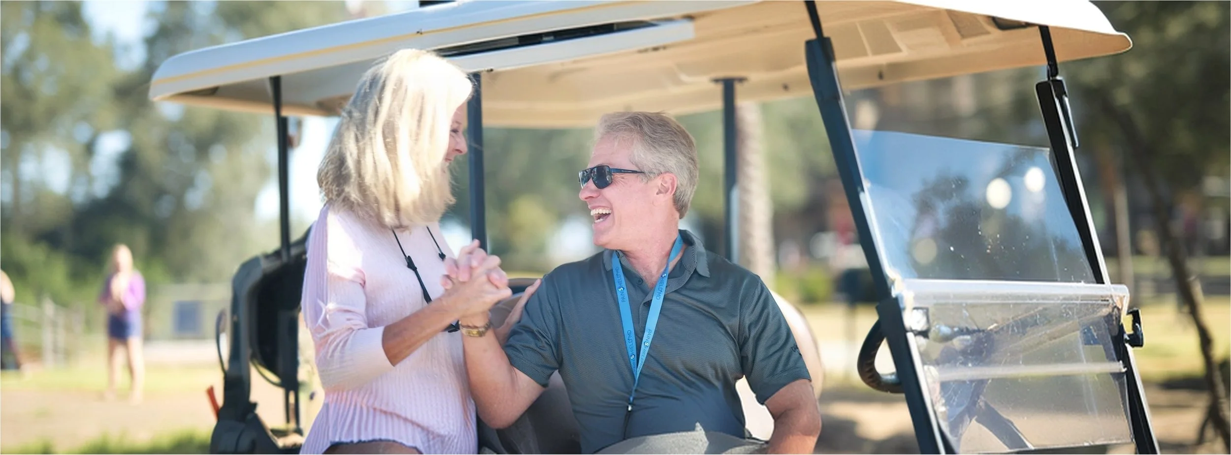 A man and woman sitting in a golf cart, smiling and holding hands, outdoors on a sunny day with trees and other people in the background.