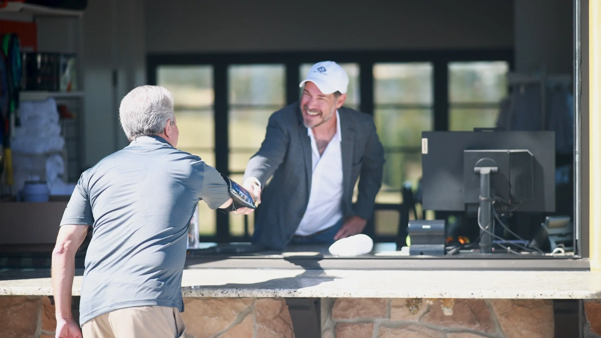 A man in a white cap and a business suit shaking hands with another man in a gray polo shirt at a counter outside a building.