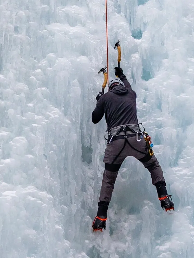 A person ice climbing on a frozen waterfall or ice wall, wearing winter gear, harness, and crampons.