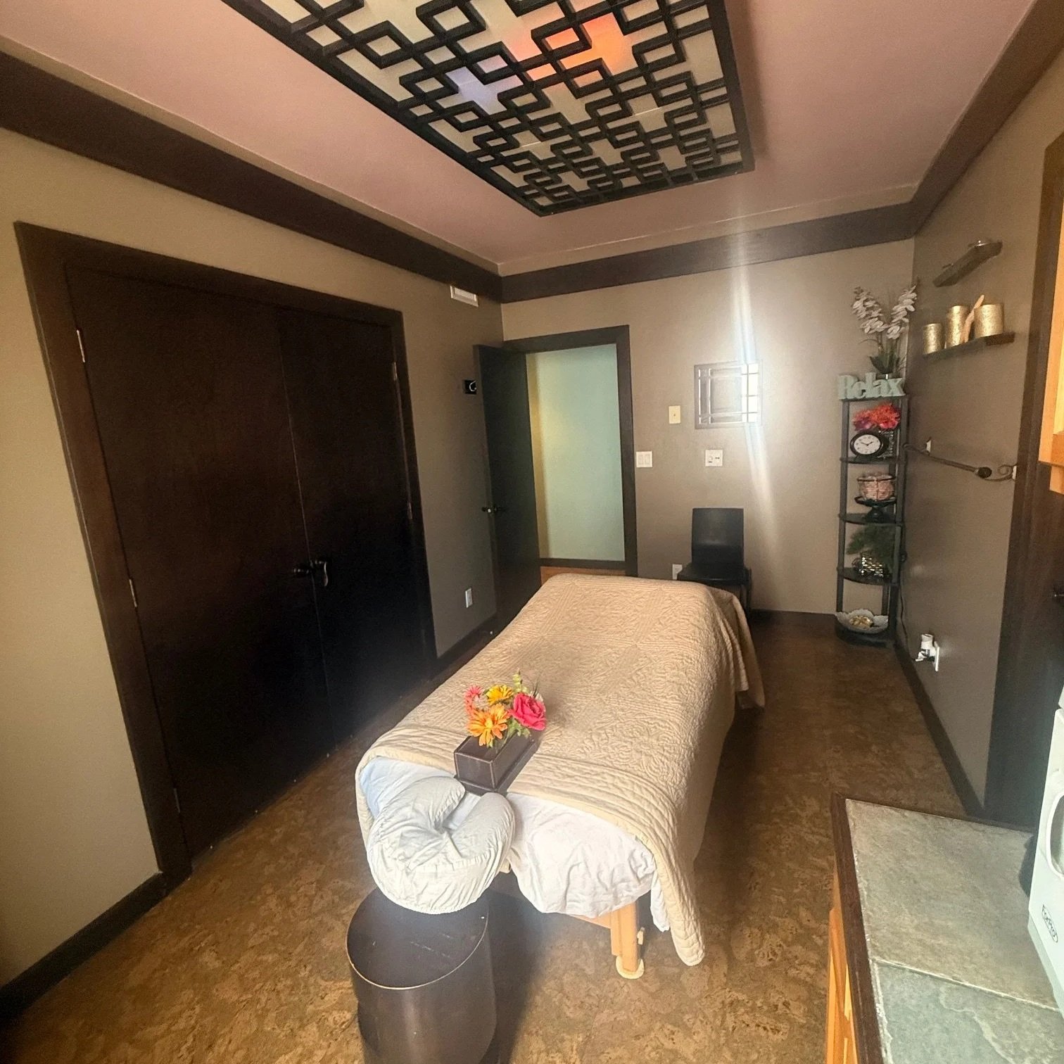A massage room with a massage table covered with a beige quilt, a small black stool with a flower arrangement on top at the foot of the table, a black chair in the corner, and a shelving unit with decorative items and plants. There is a door, a closet, and a ceiling light fixture.