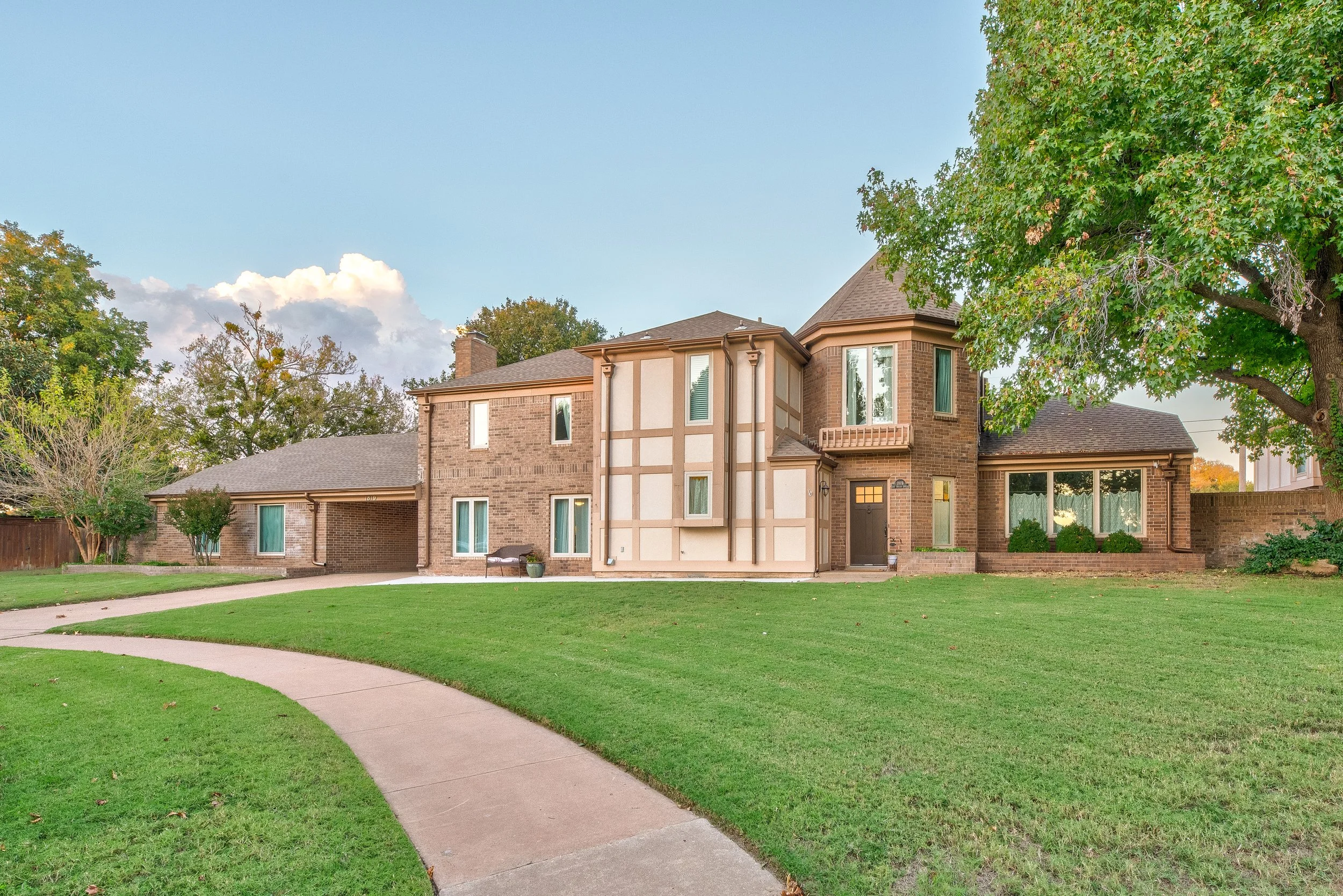 A two-story brick house with a tan and white accent in a suburban neighborhood. The house has multiple large windows, a brown front door, and a small balcony. The yard features a well-maintained green lawn, some small trees, and a concrete walkway leading to the front entrance. A large tree is visible on the right side of the image.