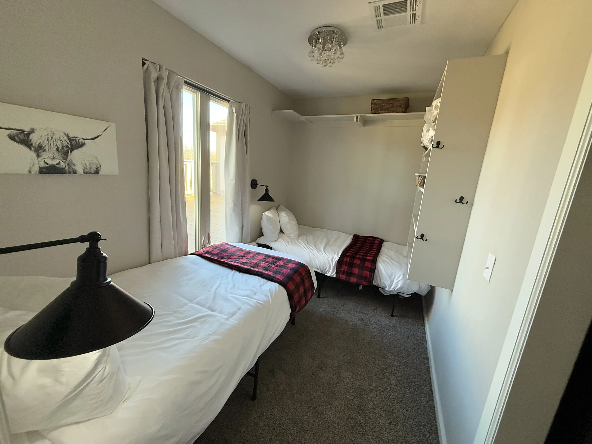 A bedroom with two beds, decorated in neutral tones, with a sliding glass door leading to a balcony, a black bedside lamp, a wall art of a cow, and a white bookshelf with hooks on the side.