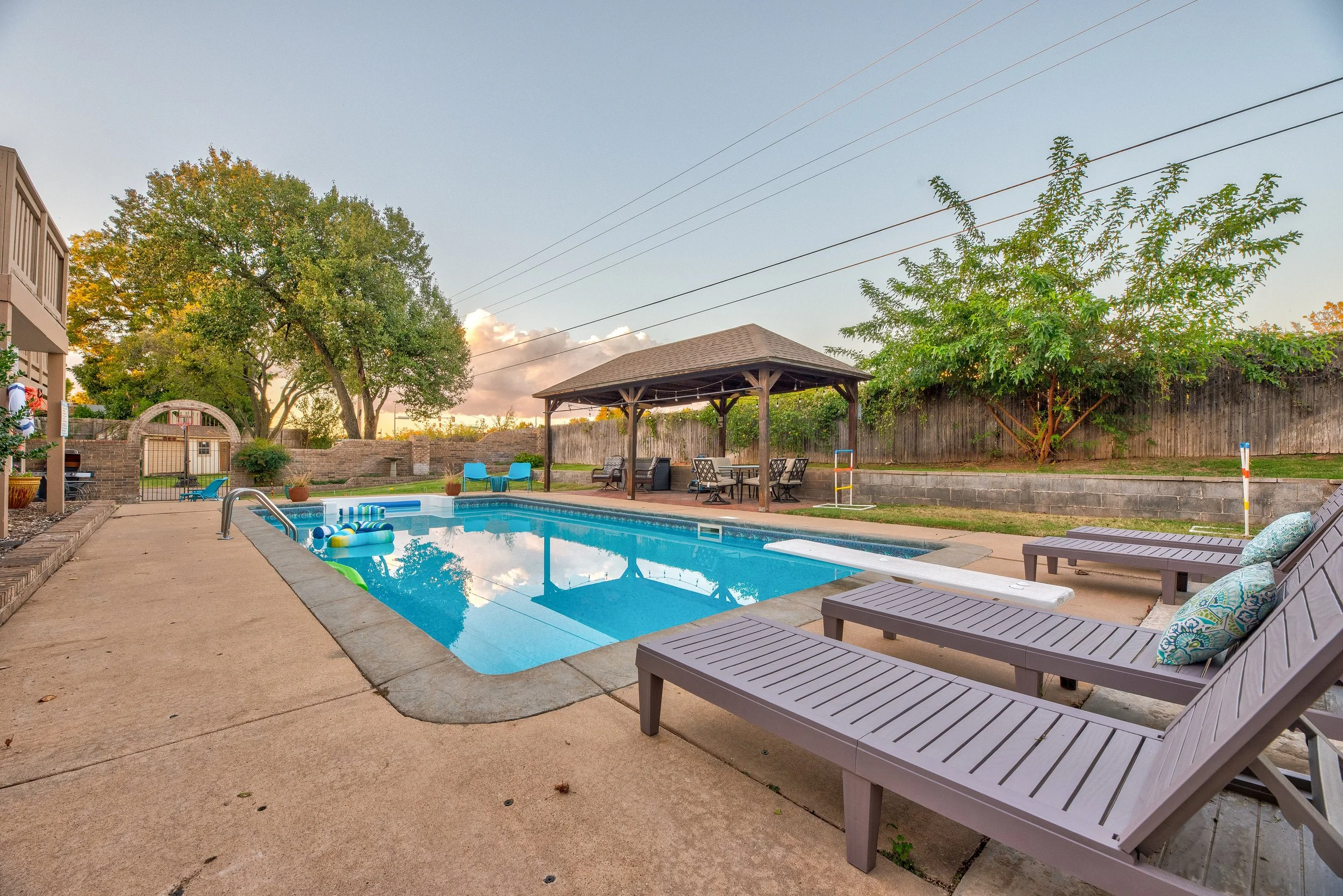 Backyard swimming pool with loungers, a shaded seating area, and trees in the background during late afternoon or early evening.