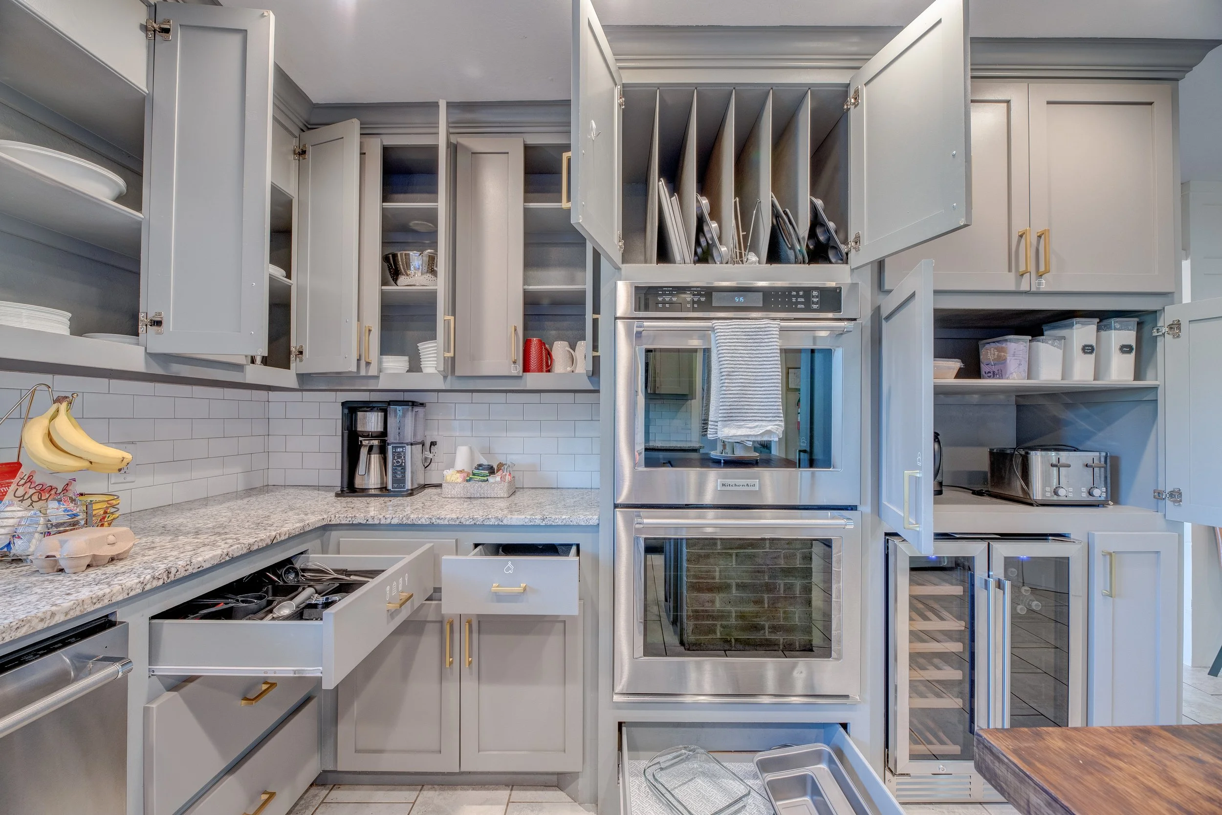 Kitchen with open cabinets, a granite countertop, a coffee maker, and a built-in oven with a towel hanging on it. Cabinets are light gray with gold handles. Open cabinet doors reveal dishes, bowls, and kitchen tools. There are bananas and a thank you