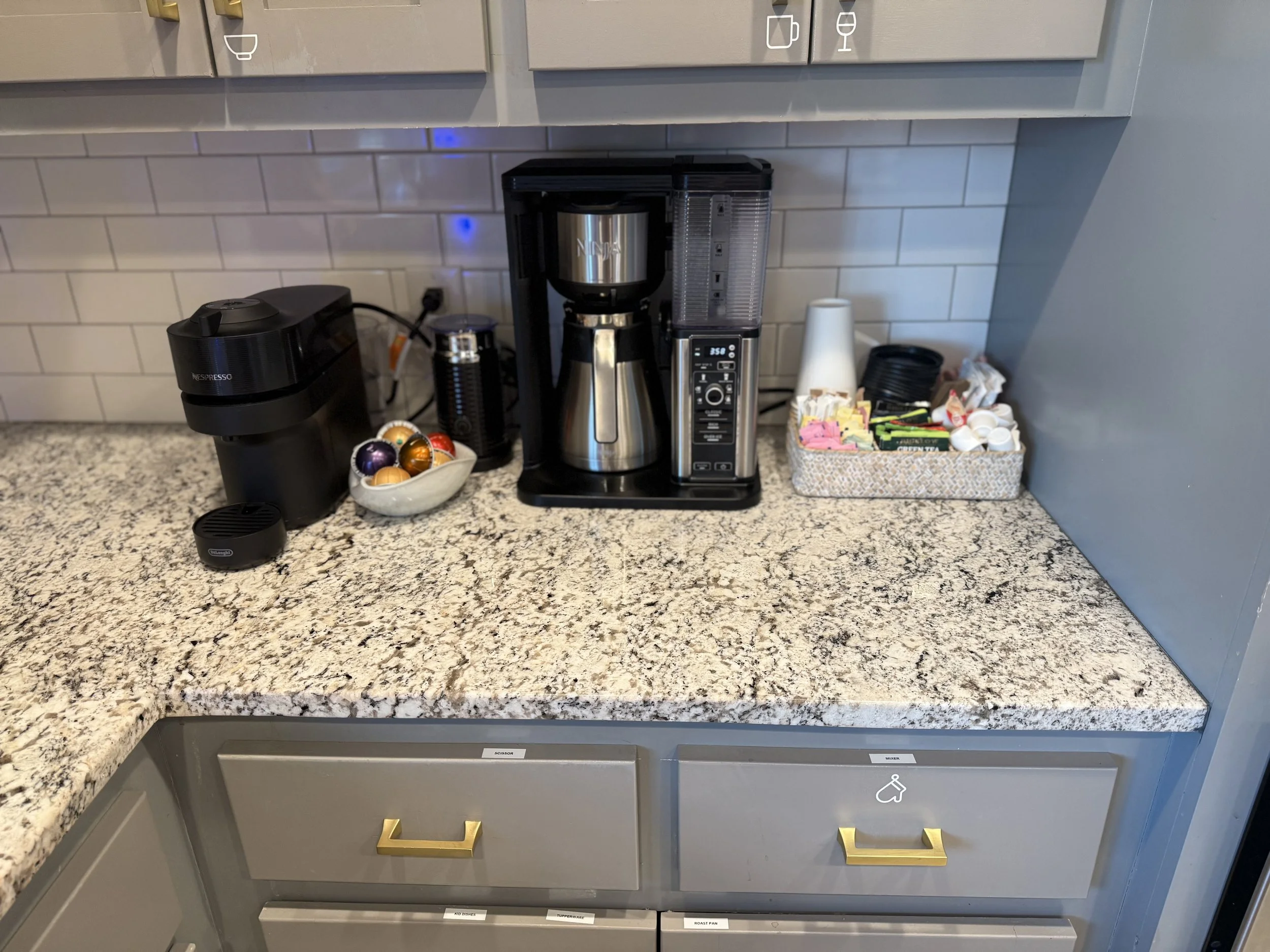 A kitchen counter with a coffee maker, a bowl of colorful coffee capsules, a small black sealed container, and a box filled with various tea bags, sugar packets, and disposable cups. There are gray cabinets with gold handles and white subway tiles on