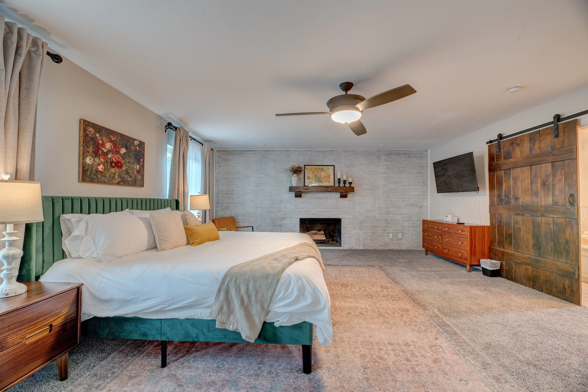 A cozy bedroom with a large bed with white bedding, a green velvet headboard, and pillows, flanked by nightstands with lamps. There is a painted floral artwork on the wall, and a white brick fireplace with a wooden mantel decorated with candles and a