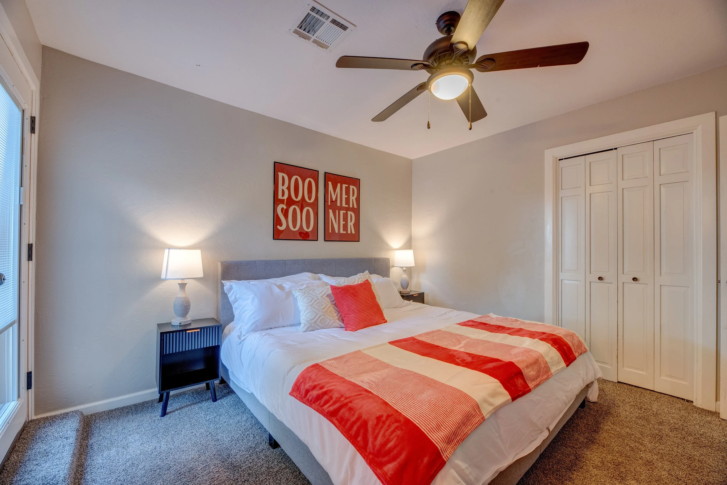 A bedroom with a bed that has white and red bedding, two white nightstands with lamps, two red framed art pieces reading "BOO SOO" and "MER NER", a ceiling fan, and a closet with white doors.