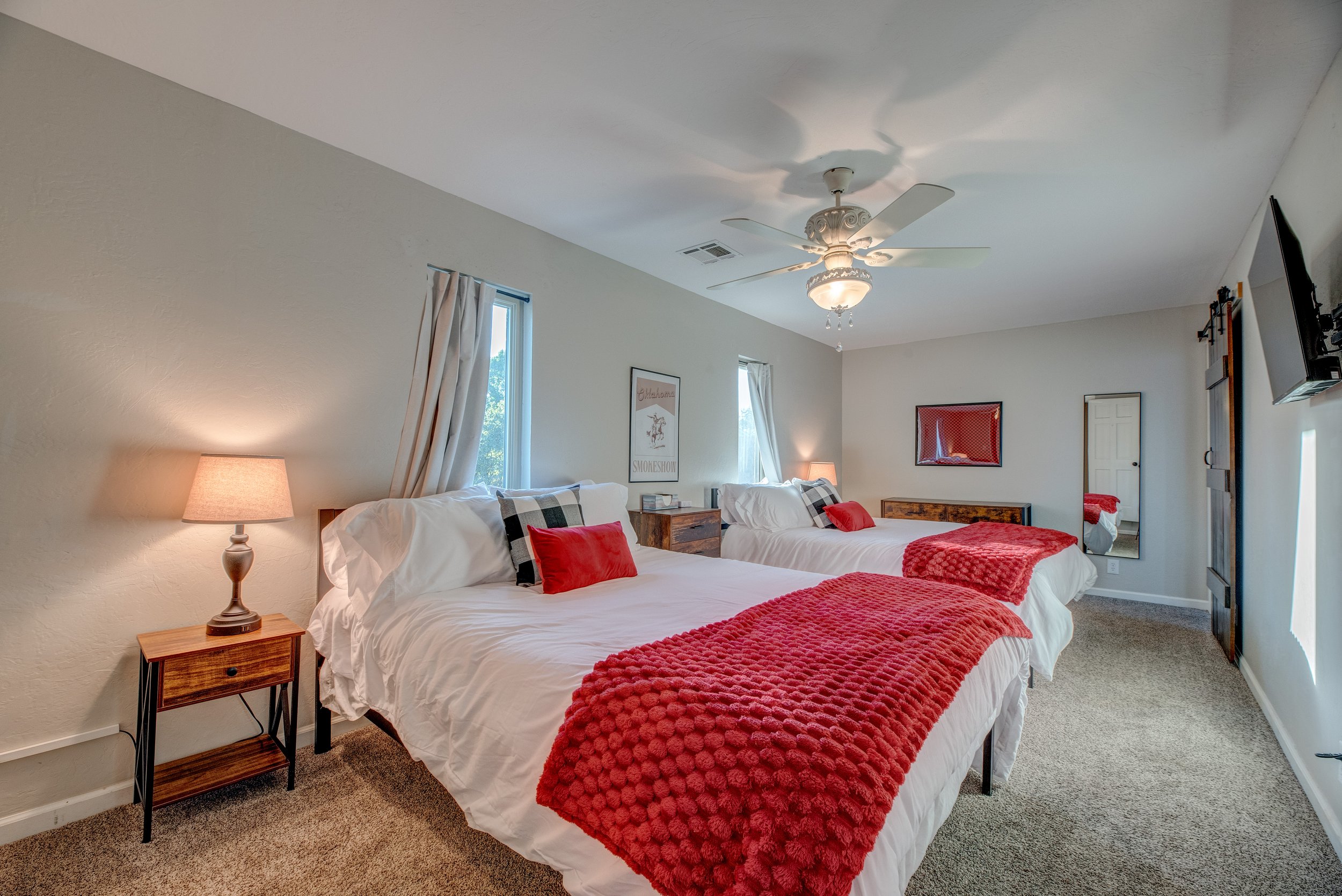 A cozy bedroom with two beds covered in white linens and red textured throws, accented with red and plaid pillows. Nightstands with lamps flank the beds. The room has a ceiling fan with light, a wall-mounted TV, framed artwork, a mirror, and windows 