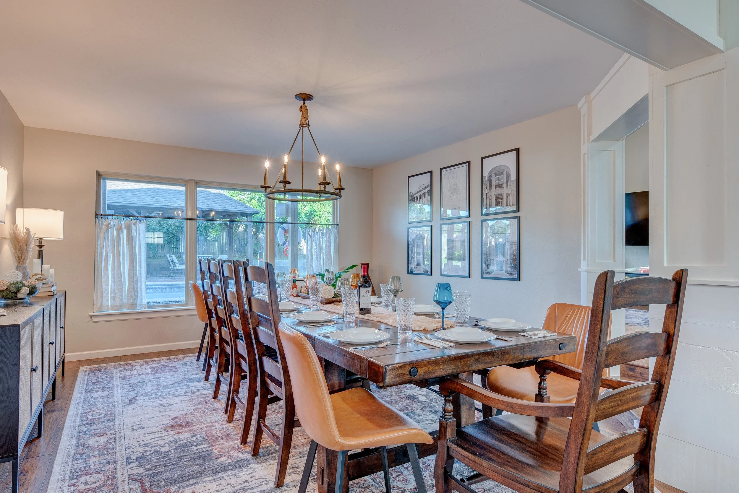A dining room with a long wooden table set for a meal, featuring glassware, plates, and a bottle of wine, with a large window showing a backyard and a chandelier hanging from the ceiling.