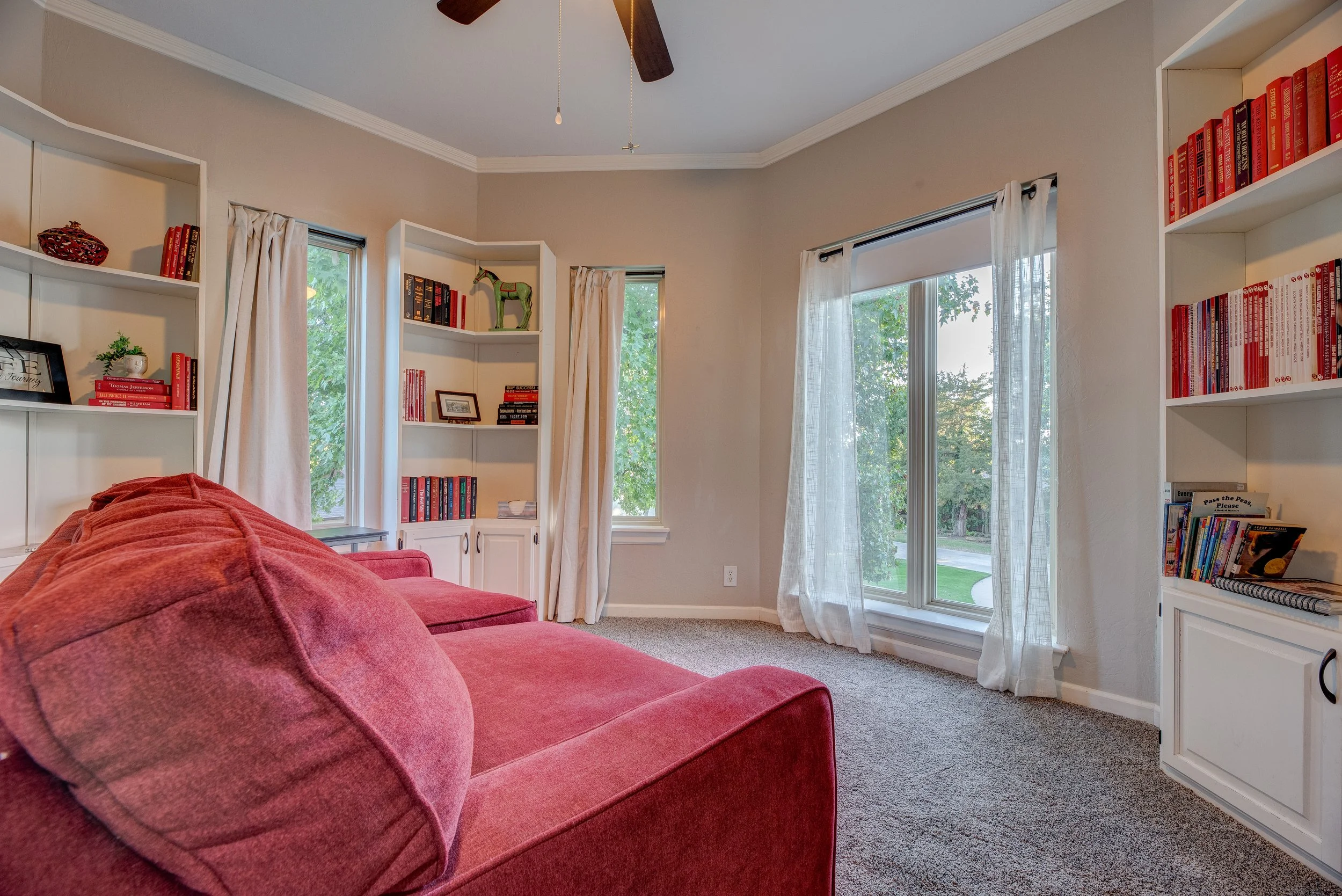 A cozy living room with beige walls, a red sofa, and white bookshelves filled with books and decorative items. Large windows with white curtains reveal green trees outside, and a ceiling fan is mounted on the ceiling.